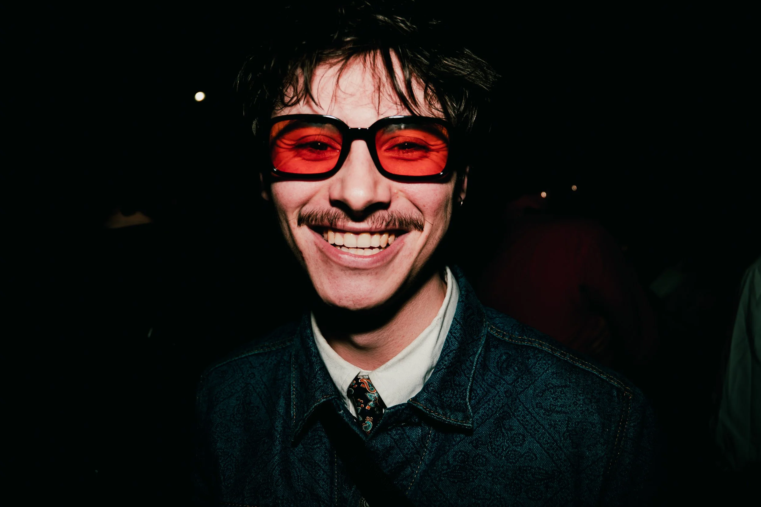 A young man with messy dark hair wearing large red-tinted glasses, a dark patterned blazer, a white shirt, and a colorful tie, smiling in a dark setting.