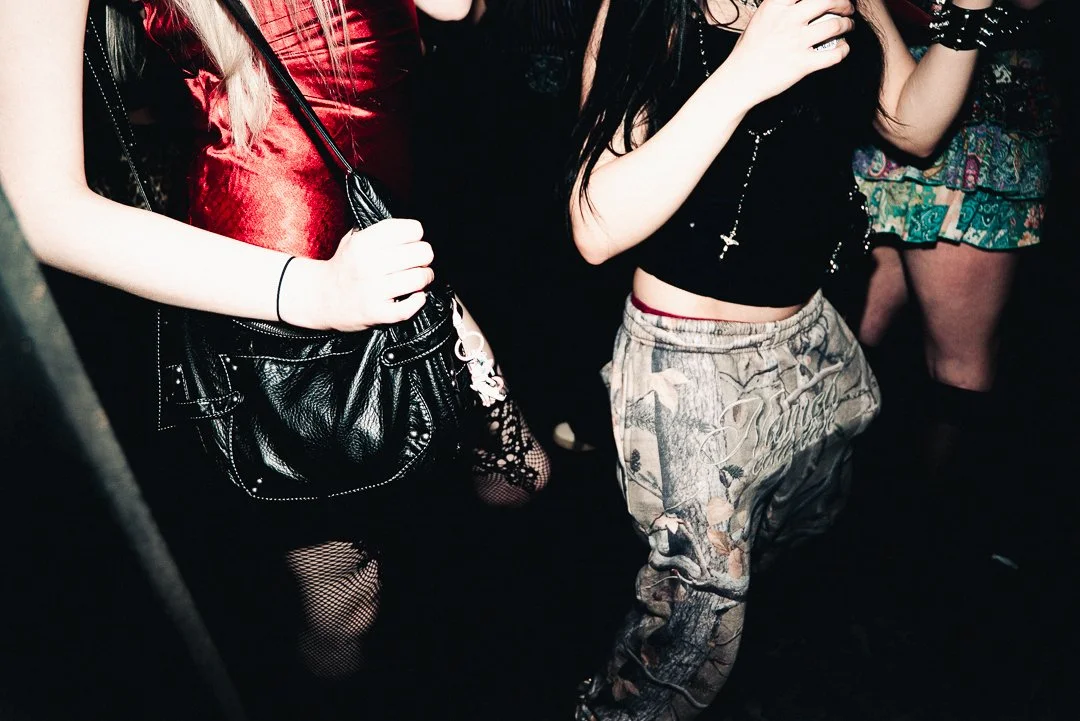 Close-up of people at a nightclub or party, showing their torsos and legs in casual and trendy outfits, holding drinks and wearing accessories.