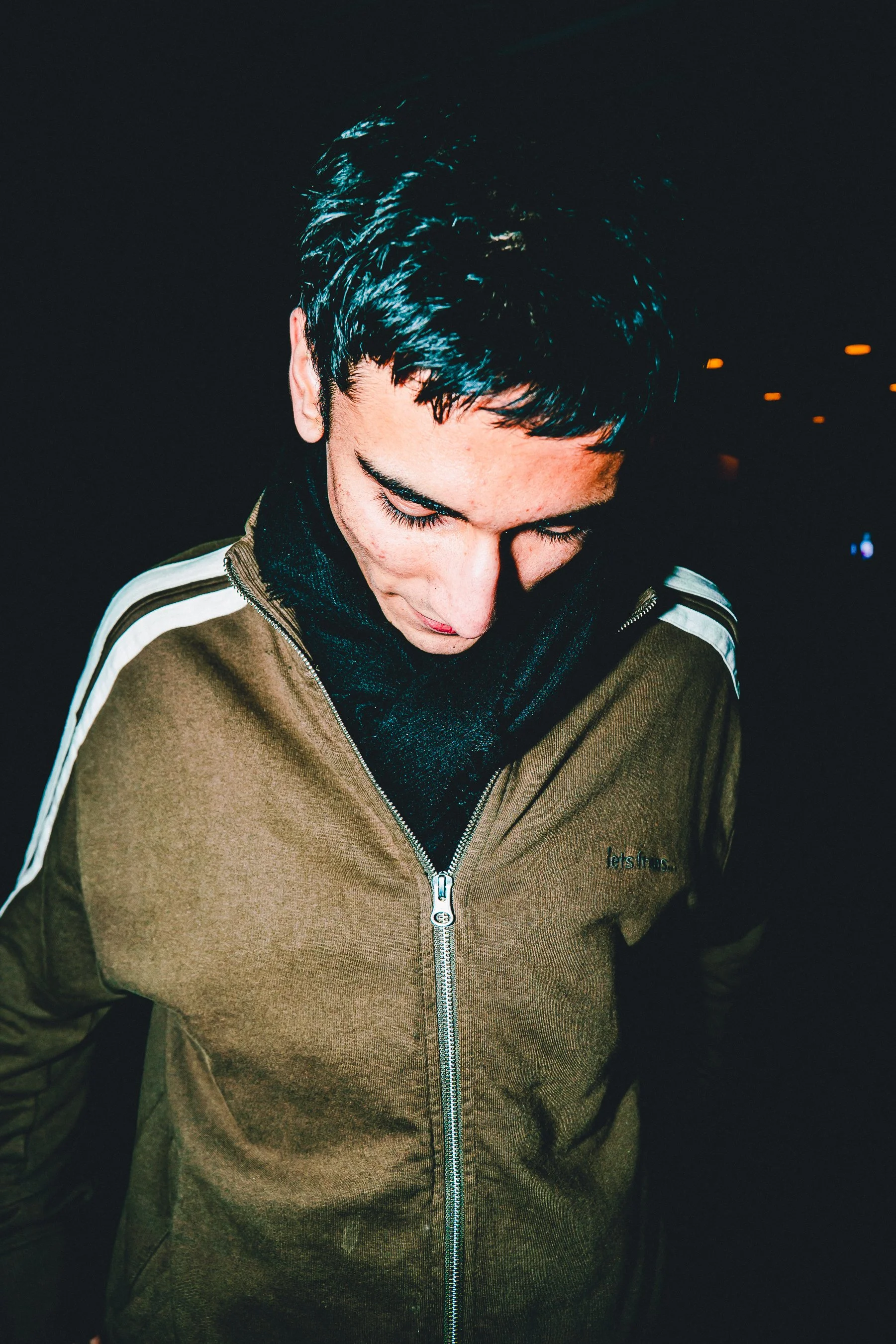 Young man with short dark hair looking down, wearing a brown zip-up jacket with white stripes on the sleeve and a black hoodie underneath, in a dark setting with small orange and blue lights in the background.