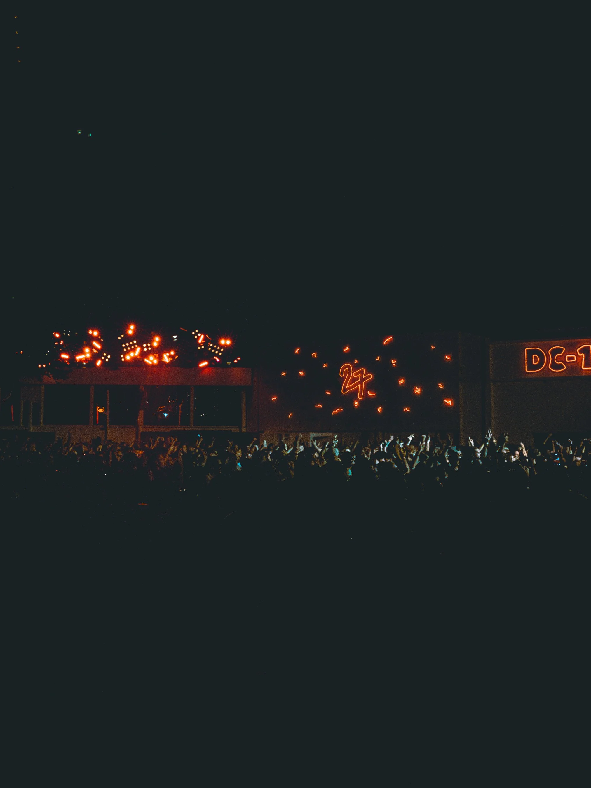 Nighttime city scene with neon signs and illuminated buildings, street lights, and a crowd of people in the foreground.