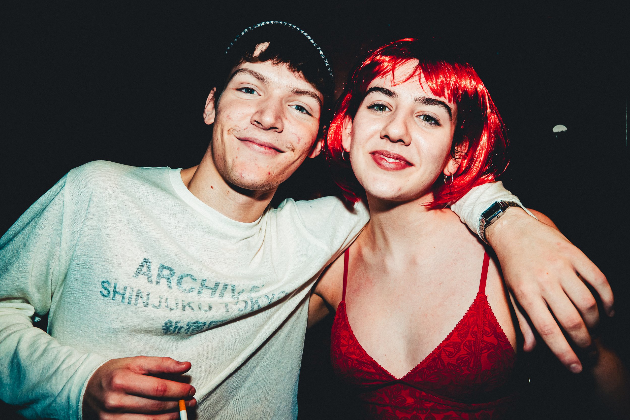 Two young adults, a man with dark hair and a woman with bright red hair, standing close together and smiling at the camera. The woman is wearing a red lace top, and the man is wearing a light-colored shirt with text on it. They are in a dark setting,