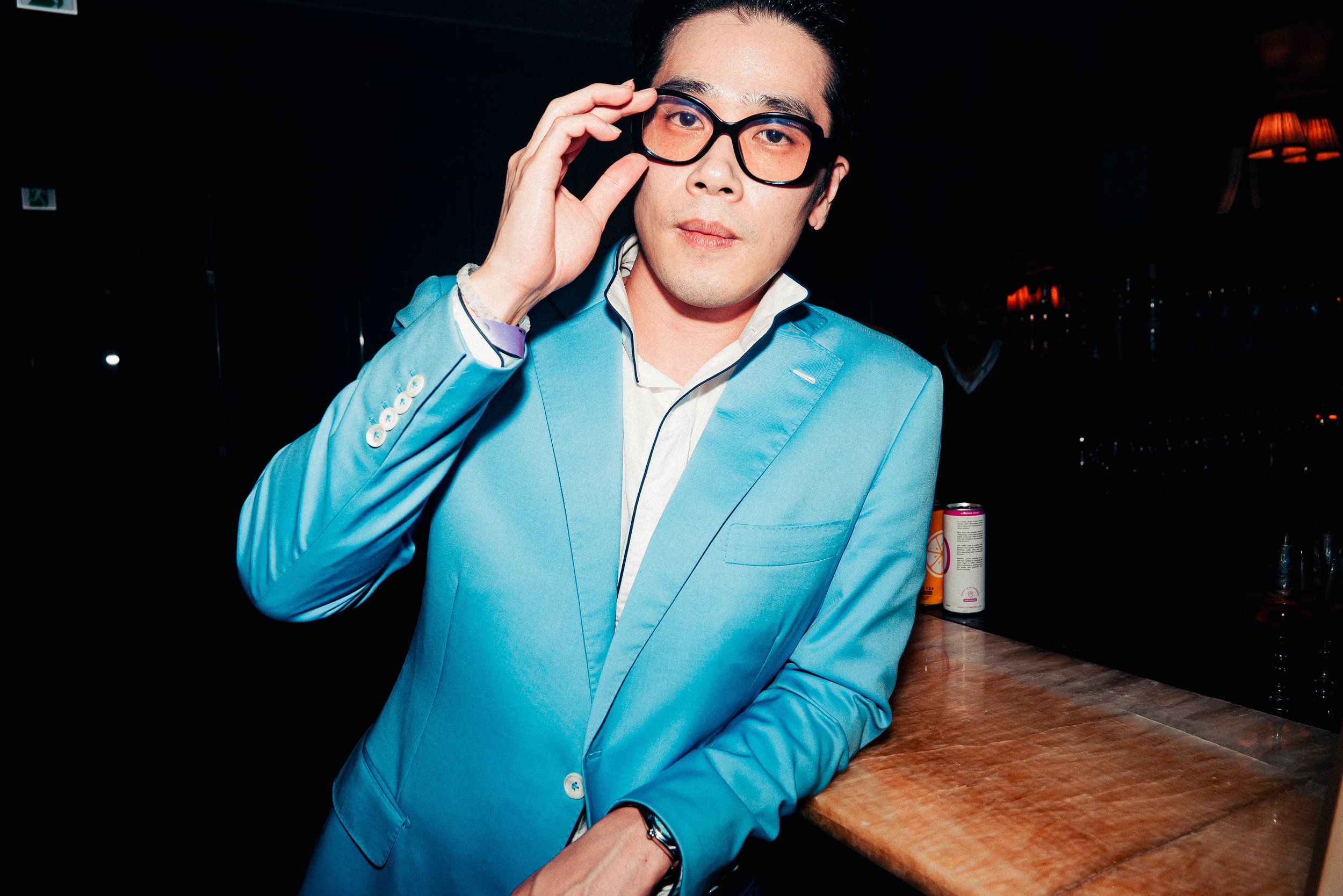 A man wearing a bright blue blazer and black glasses adjusting his glasses with his right hand, standing near a table in a dimly lit room with warm lighting.