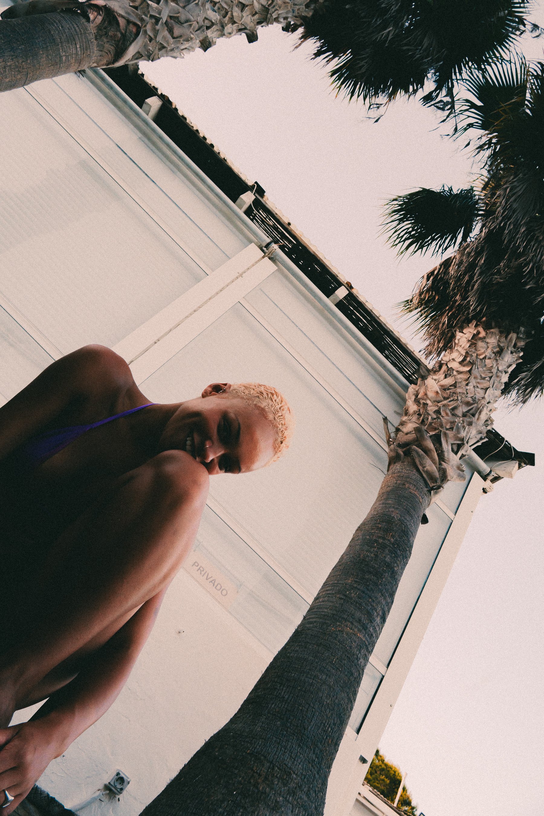 A person with platinum blonde hair smiling and sitting outdoors near a palm tree, with a white building wall and roofline in the background.