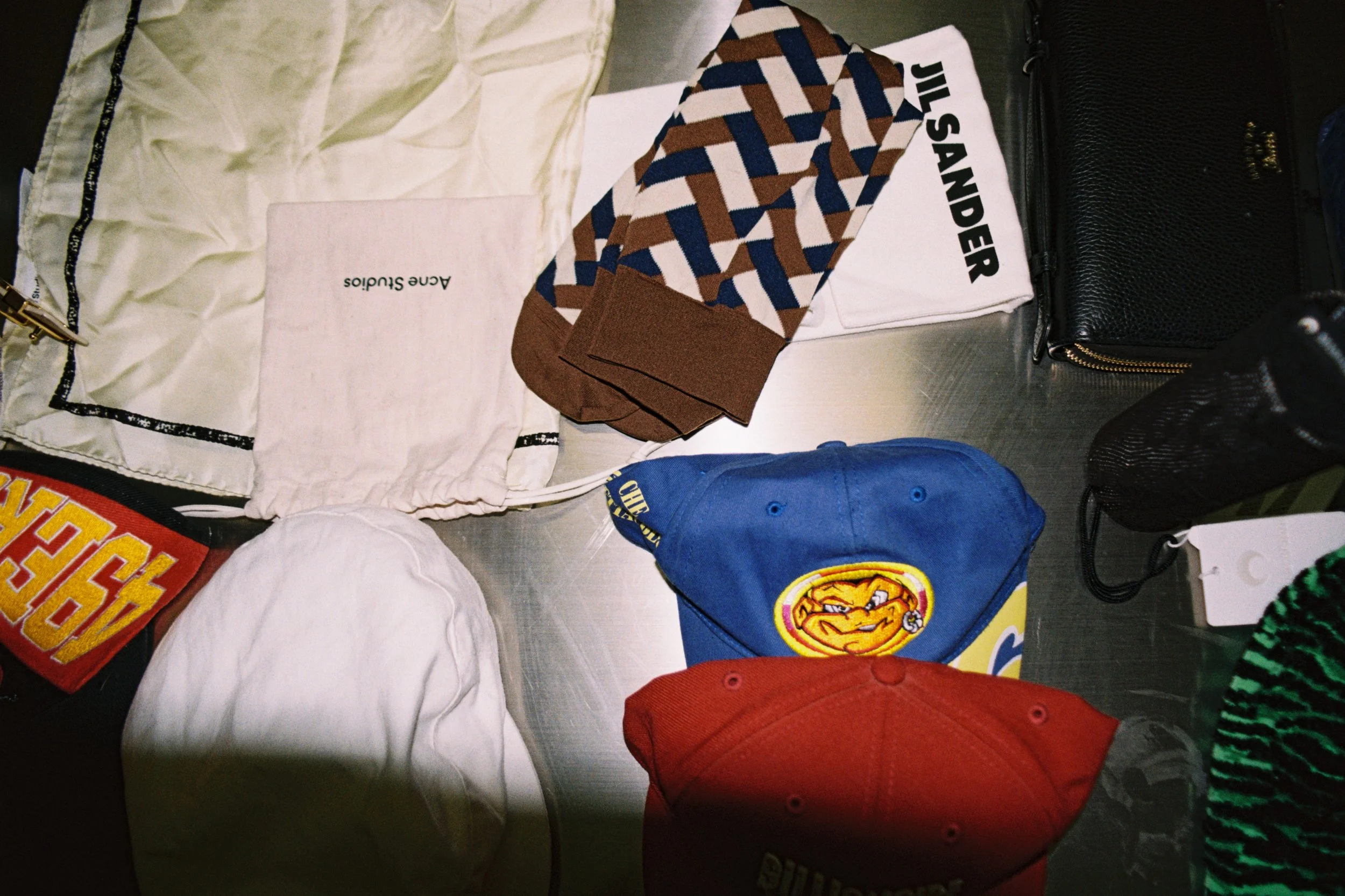 Various clothing items and accessories including a white cloth with black trim, a pair of patterned socks, a black textured bag, a blue cap with a logo, and a red and green patterned hat, all placed on a flat surface.