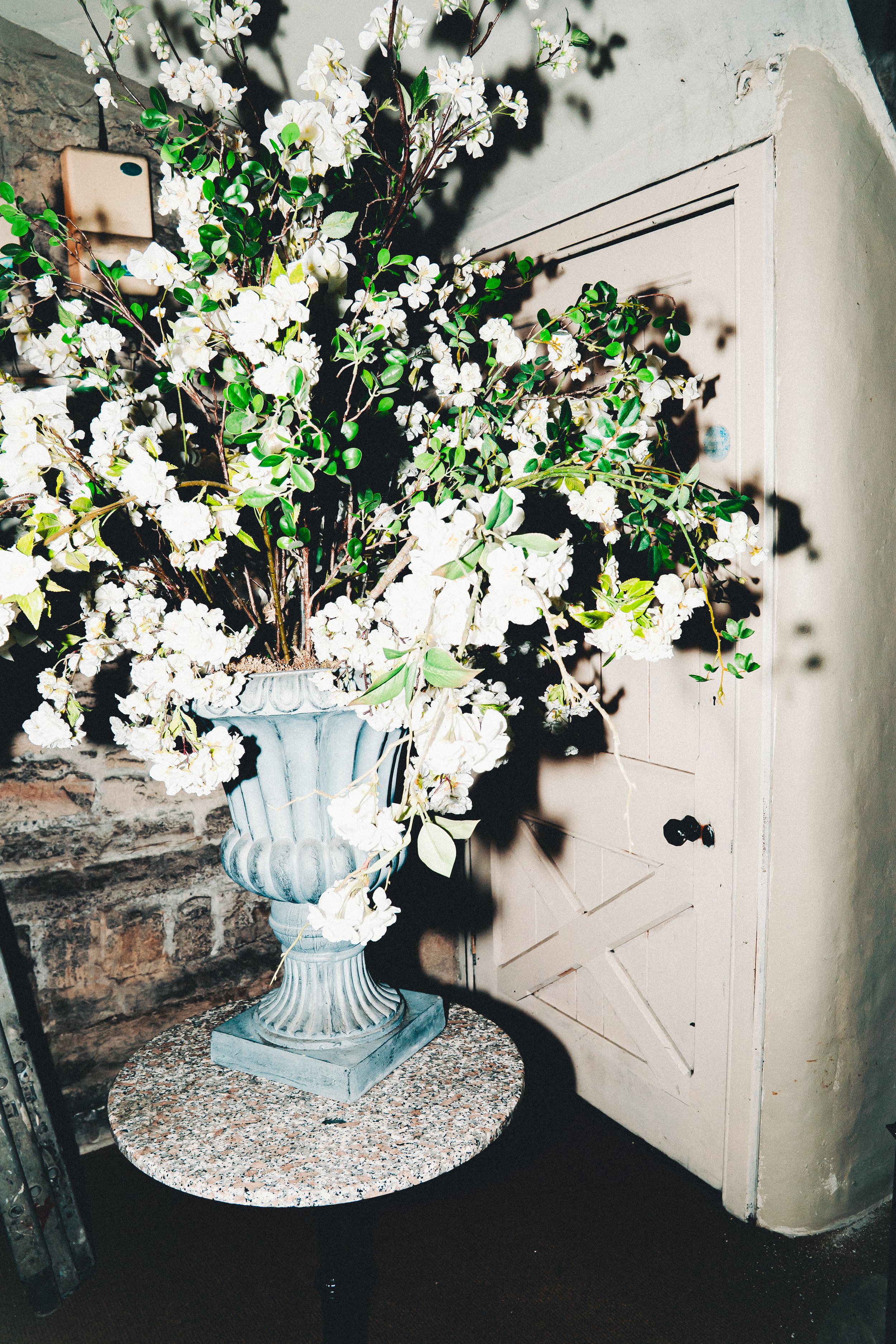 A decorative flower arrangement in a blue vase placed on a round granite table against a brick wall and a white wooden door.