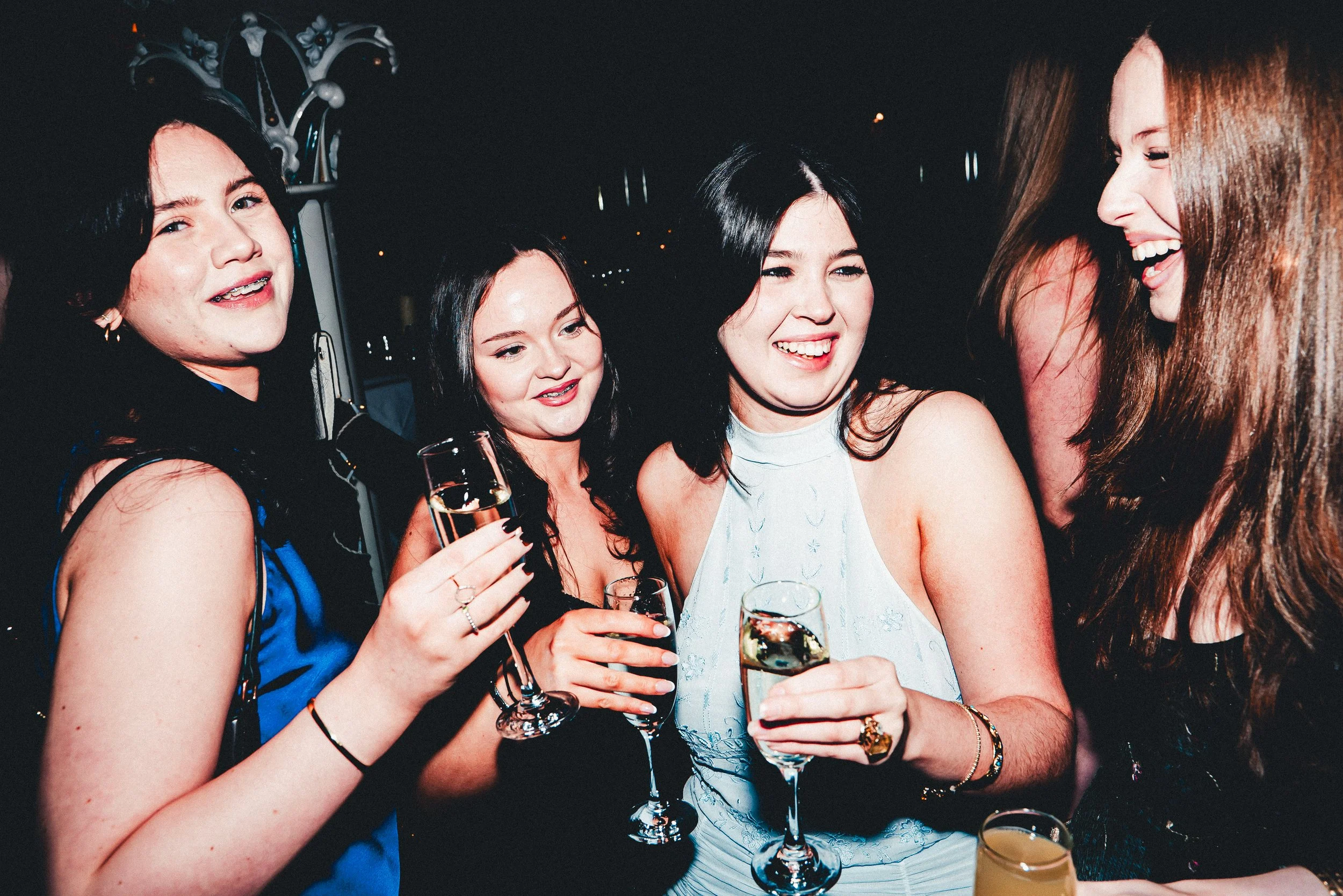 A group of four women at a party, smiling and holding glasses of champagne and orange juice, dressed in evening wear, with dark hair, in a dimly lit setting.