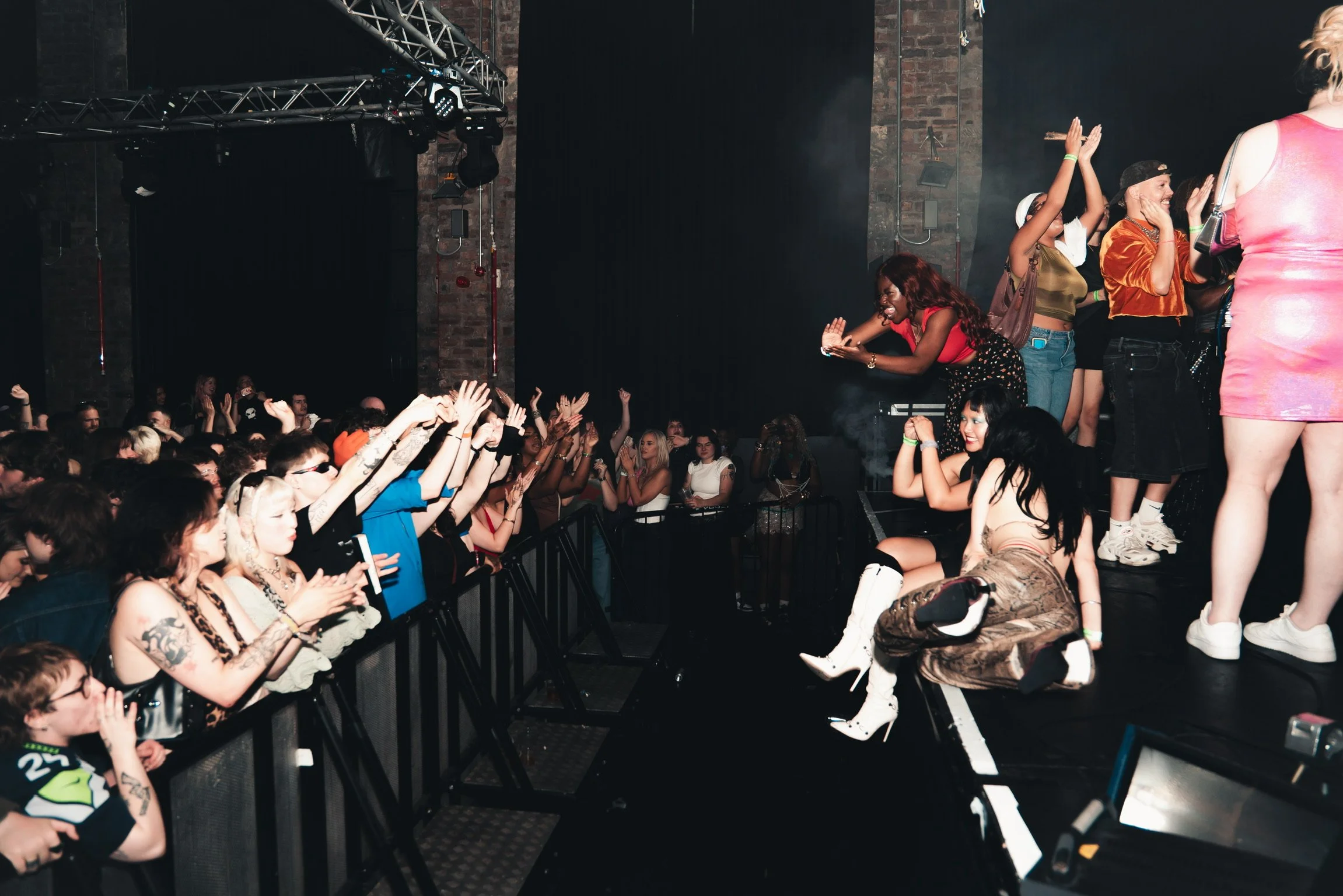 A lively concert scene with a diverse crowd of excited fans clapping and cheering, some sitting and others standing near the stage, where performers are engaging with the audience.