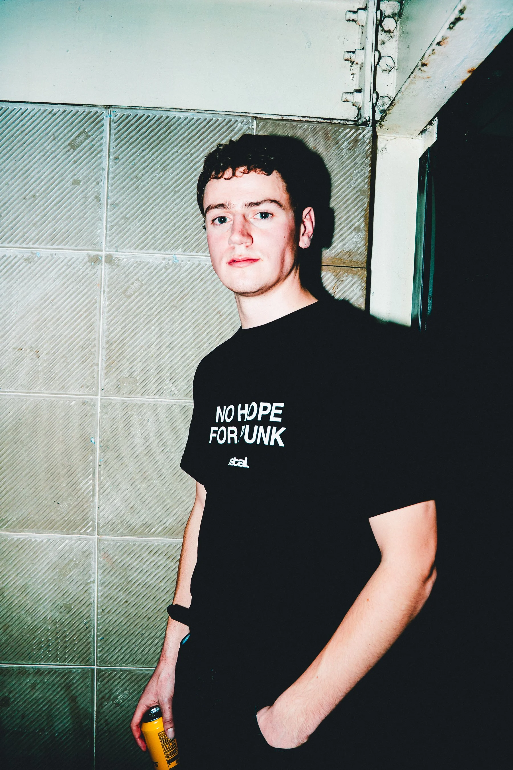 A young man with curly hair and fair skin standing indoors against a textured wall with metallic tiles. He is wearing a black t-shirt with white text, holding a yellow spray can, and looking at the camera.