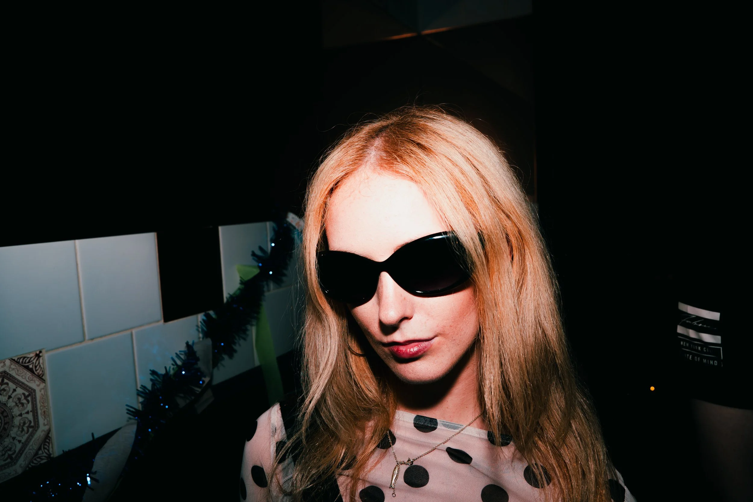 A woman with long blonde hair wearing black sunglasses and a pink top with black polka dots, indoors against a dark background with tiles and a decorated Christmas tree.