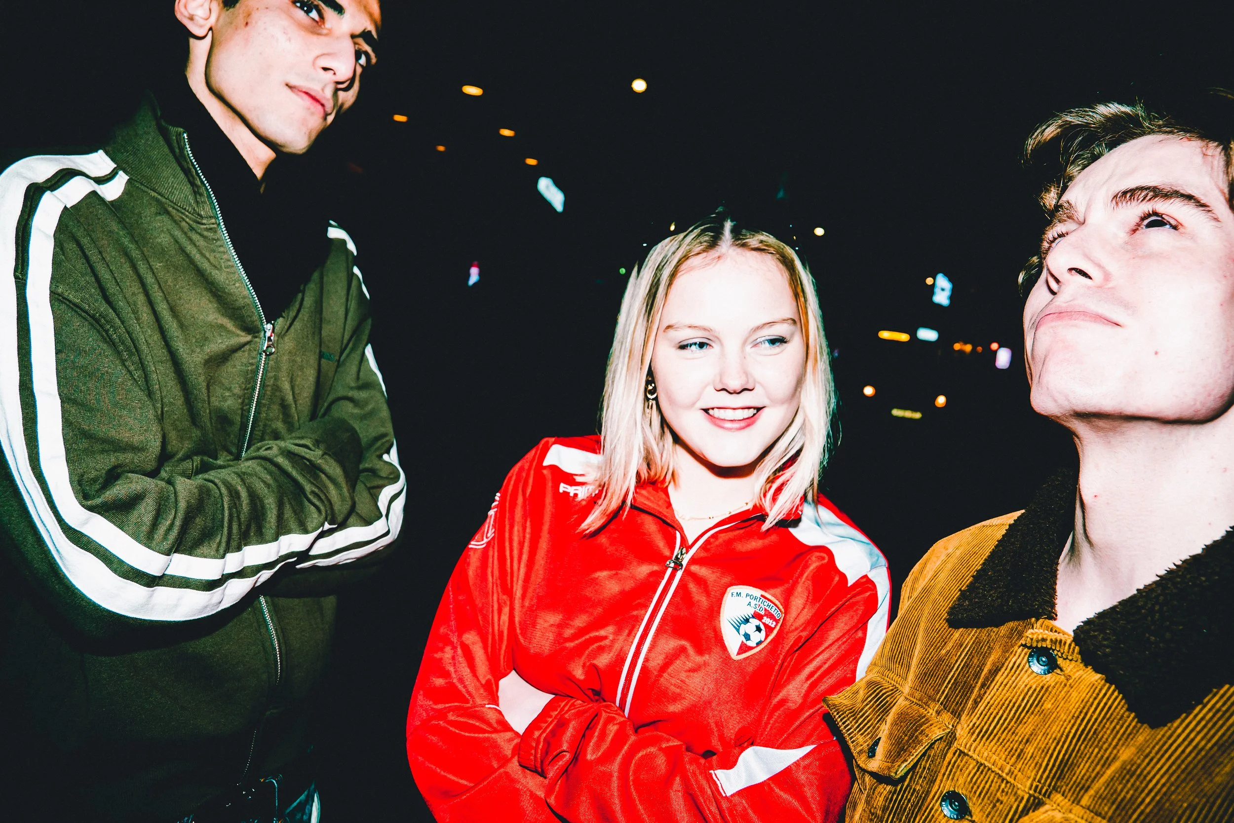 Three people standing outside at night, engaging in conversation. One man on the left is wearing a green jacket with white stripes, the woman in the middle is wearing a red sports jacket with a logo, and the man on the right is wearing a brown jacket