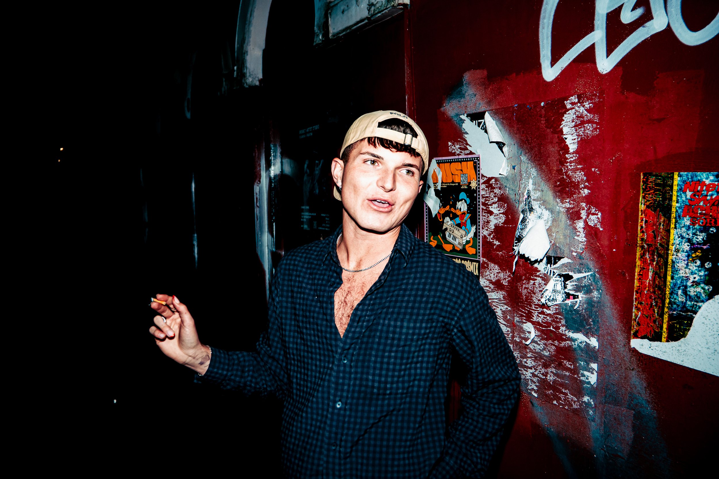 A young person with short dark hair, wearing a beige cap backwards and a dark blue checkered shirt, standing in front of a red, peeling wall with posters. The person appears to be speaking or singing, with one hand raised.