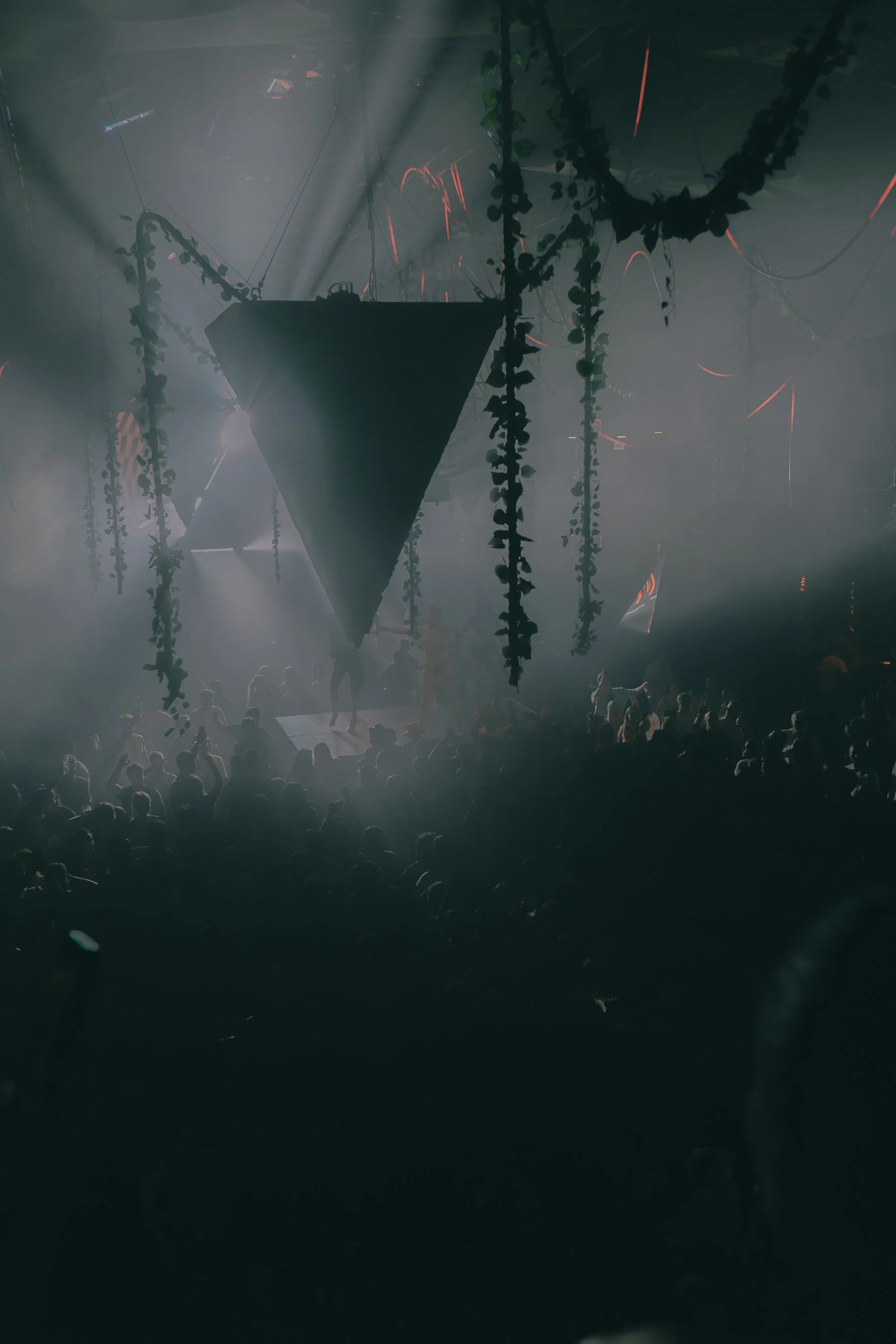 A dark concert scene with a large crowd, stage fog, hanging vines, and a triangular hanging decoration, illuminated by spotlights and red lights.