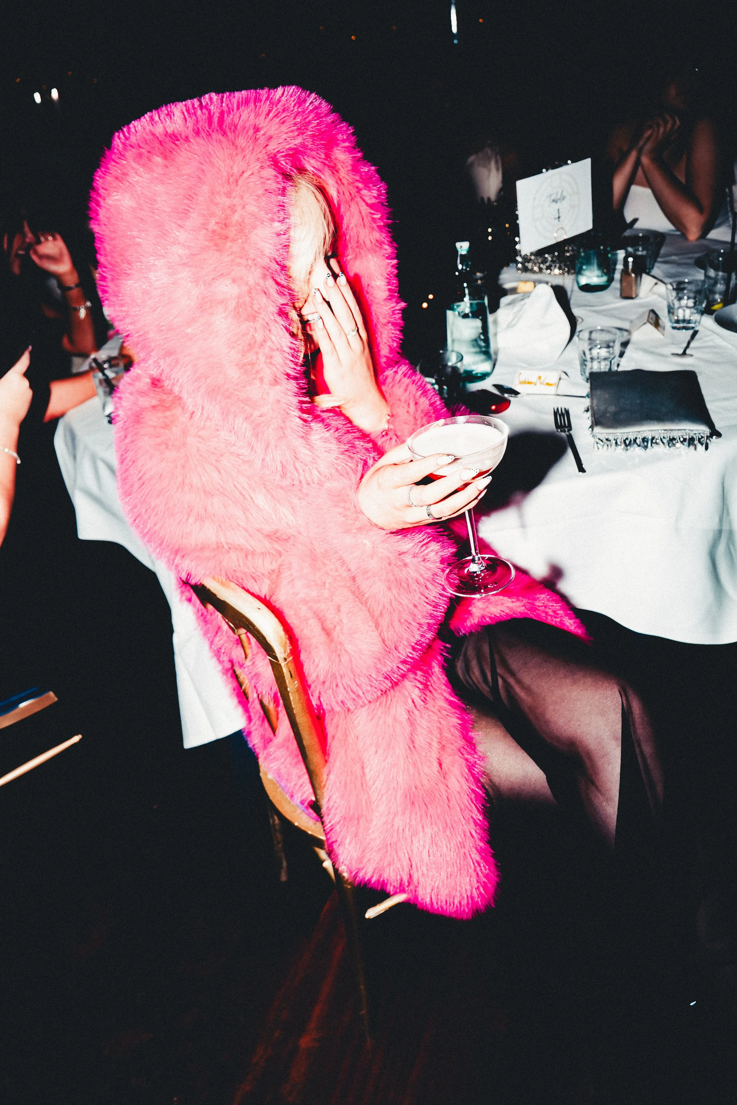 A person dressed in a bright pink fuzzy coat with a matching hood, sitting at a table with glassware, a black clutch, and a white purse in a dimly lit setting, covering part of their face with one hand and holding a glass in the other.