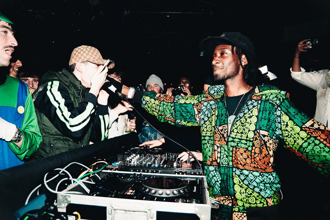 DJ wearing a colorful patterned jacket and black hat performing at a nightclub, with a crowd of young people watching and taking photos.