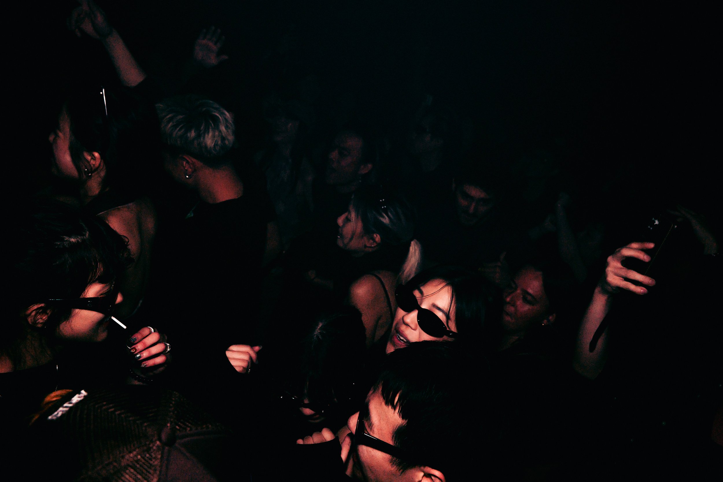 Group of people in a dark setting, many smiling and wearing sunglasses, seemingly enjoying a party or concert.