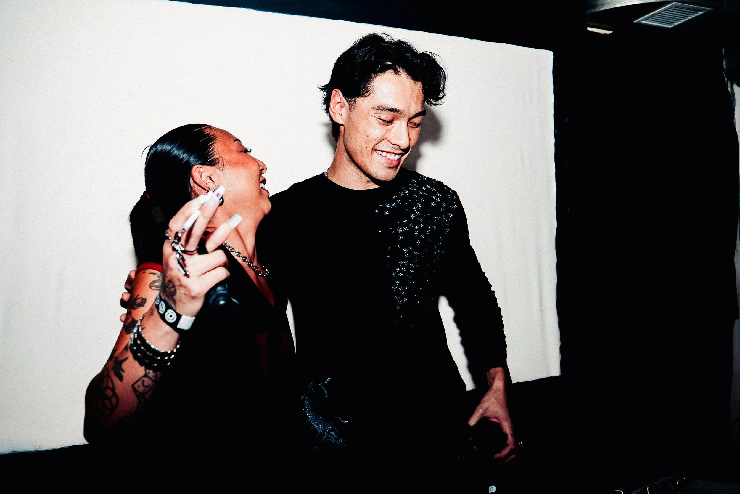 A woman with tattoos wearing jewelry and holding a cigarette walks beside a smiling man with dark hair, dressed in a black shirt with silver details, against a plain white background.