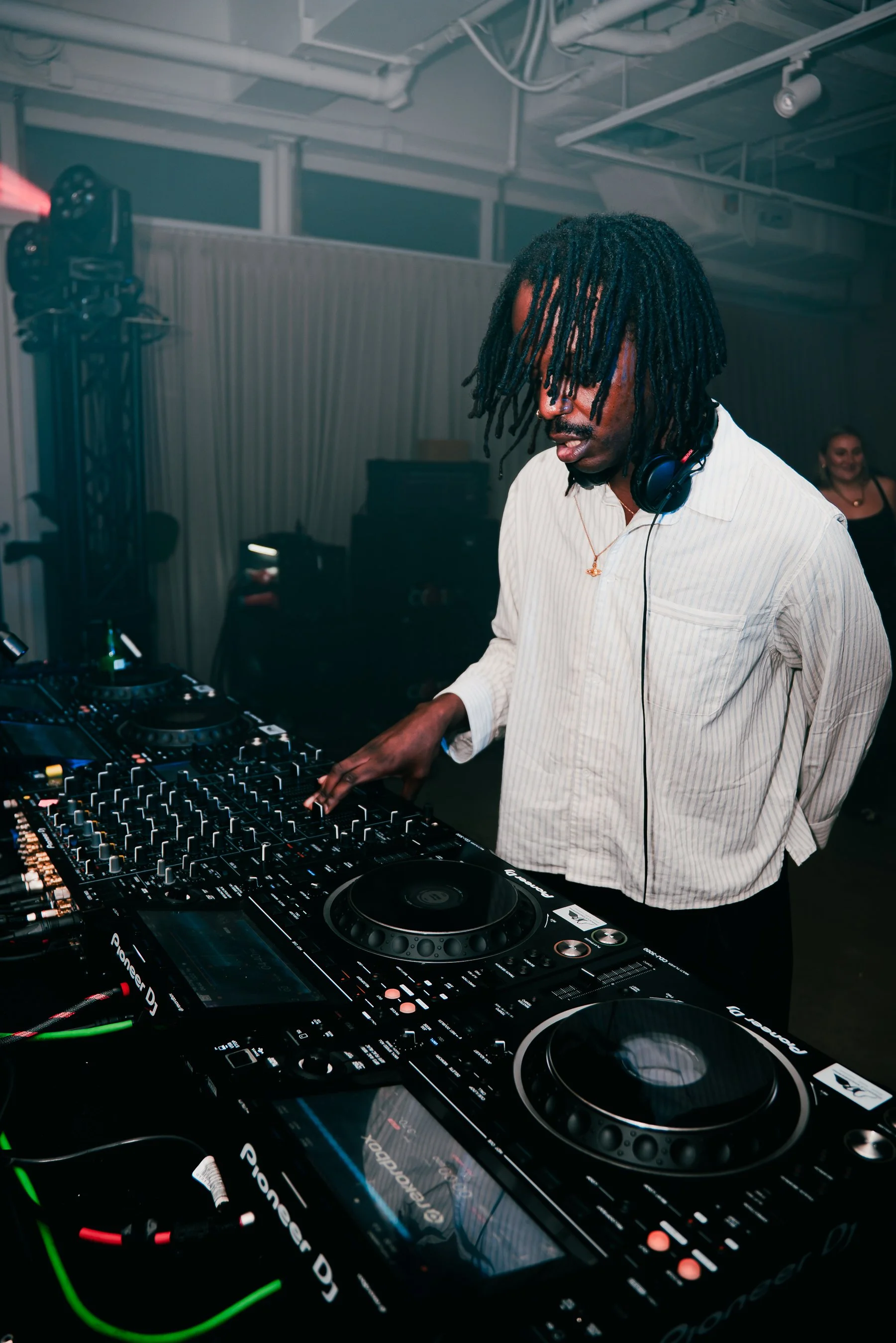A DJ with dreadlocks wearing a white shirt and headphones, operating a DJ controller at a party or event, with a woman smiling in the background.