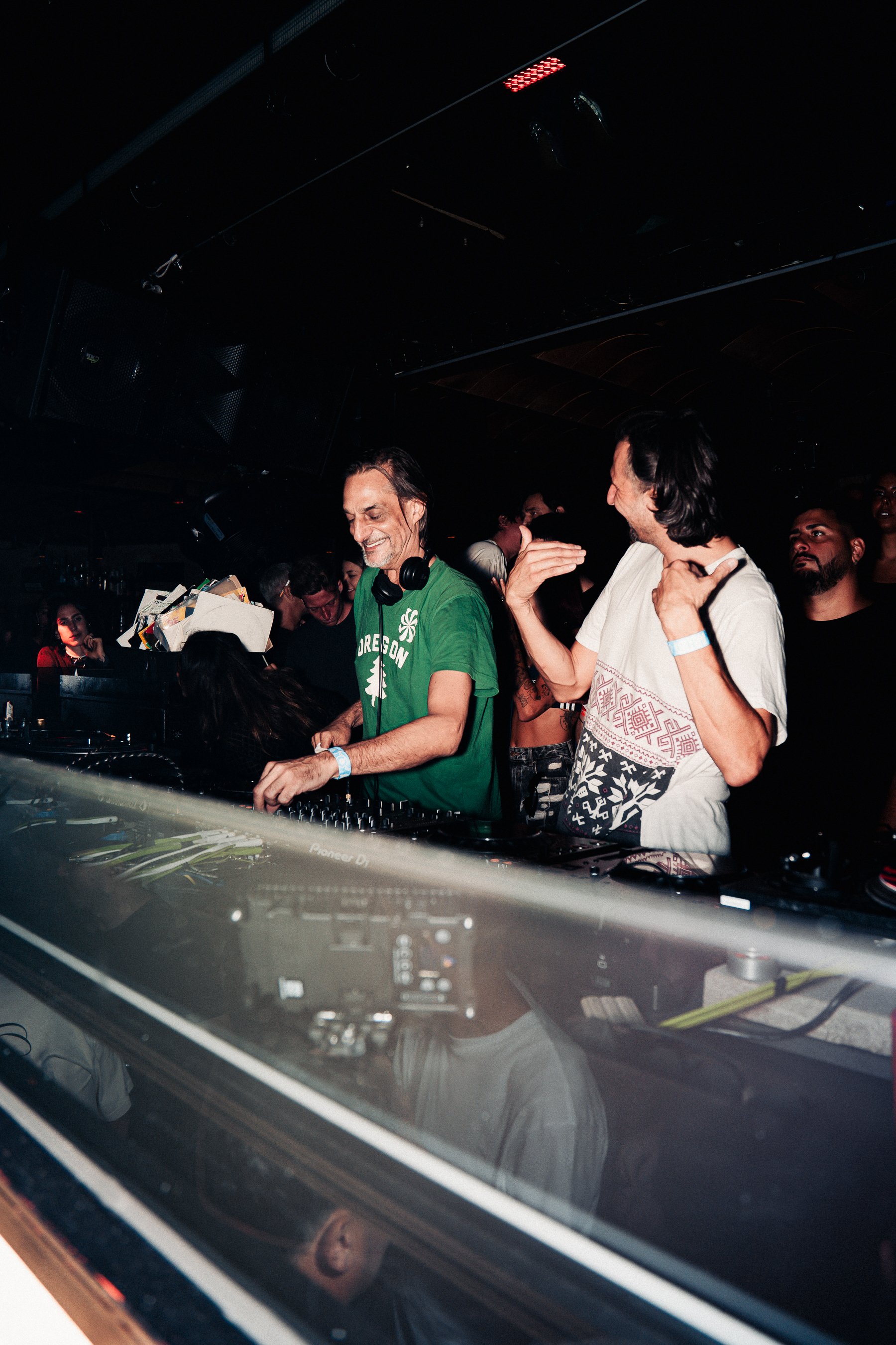 DJ in a green shirt with headphones around neck, smiling, playing music at nightclub with crowd around, seen through a DJ booth glass.