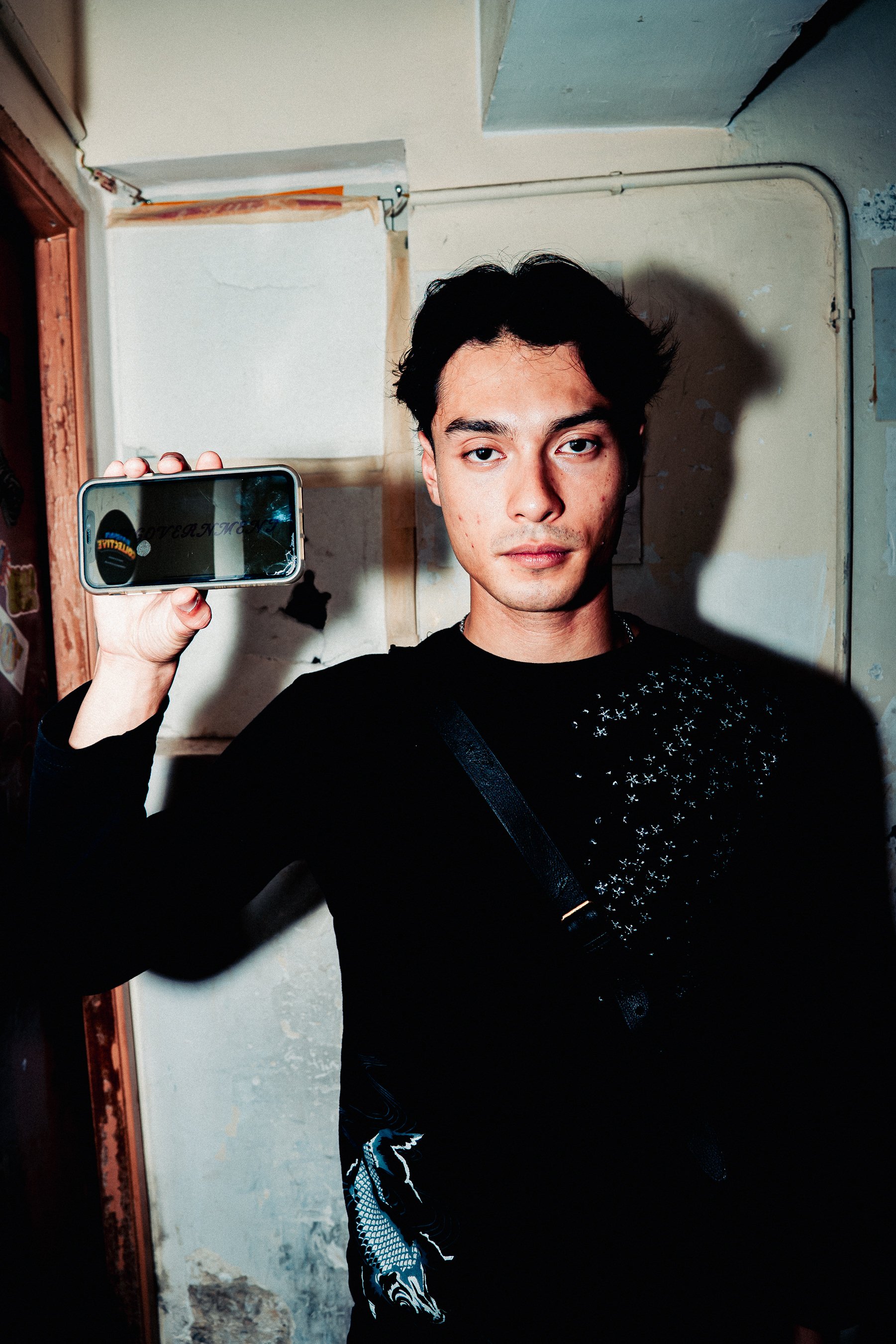 Young man with dark hair and a slight smile taking a mirror selfie with a smartphone in a room with peeling paint and exposed wiring.