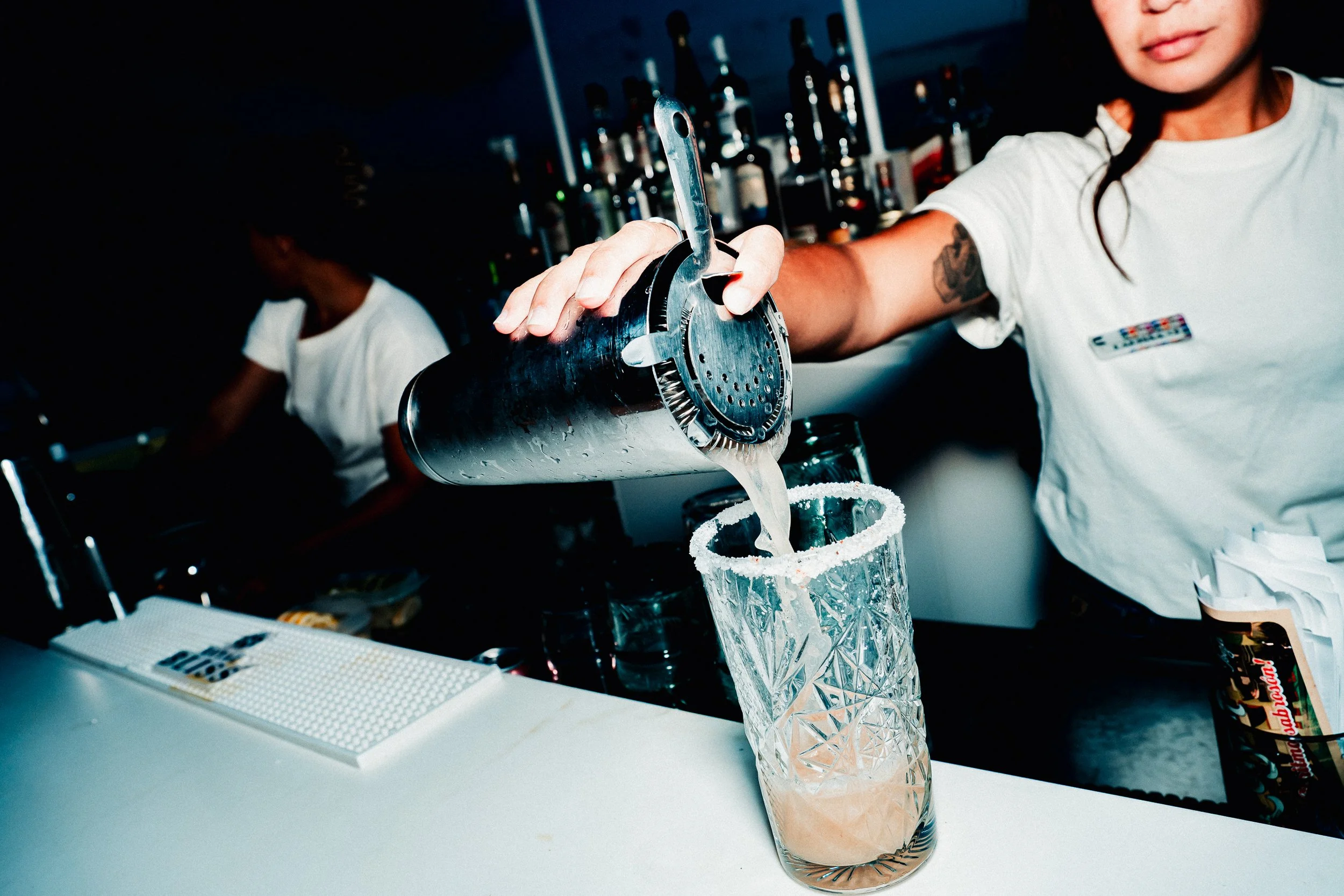 A woman pouring a drink into a tall glass with salt on the rim, in a setting with bottles on shelves in the background.