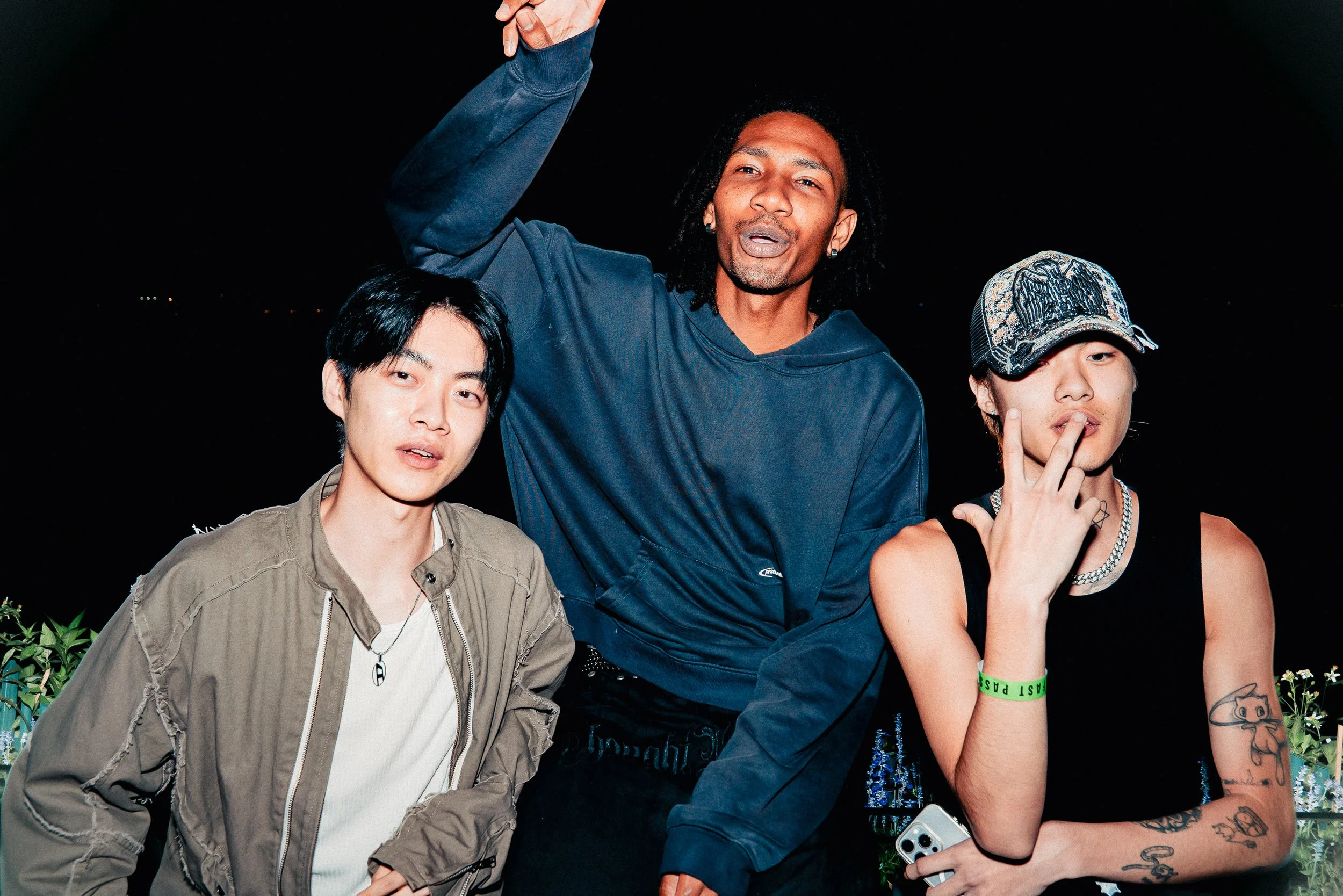 Three young adults posing together at night outdoors, with one man in a blue hoodie raising his arm, and two women, one with tattoos and wearing a cap and jewelry, all making signs or gestures.