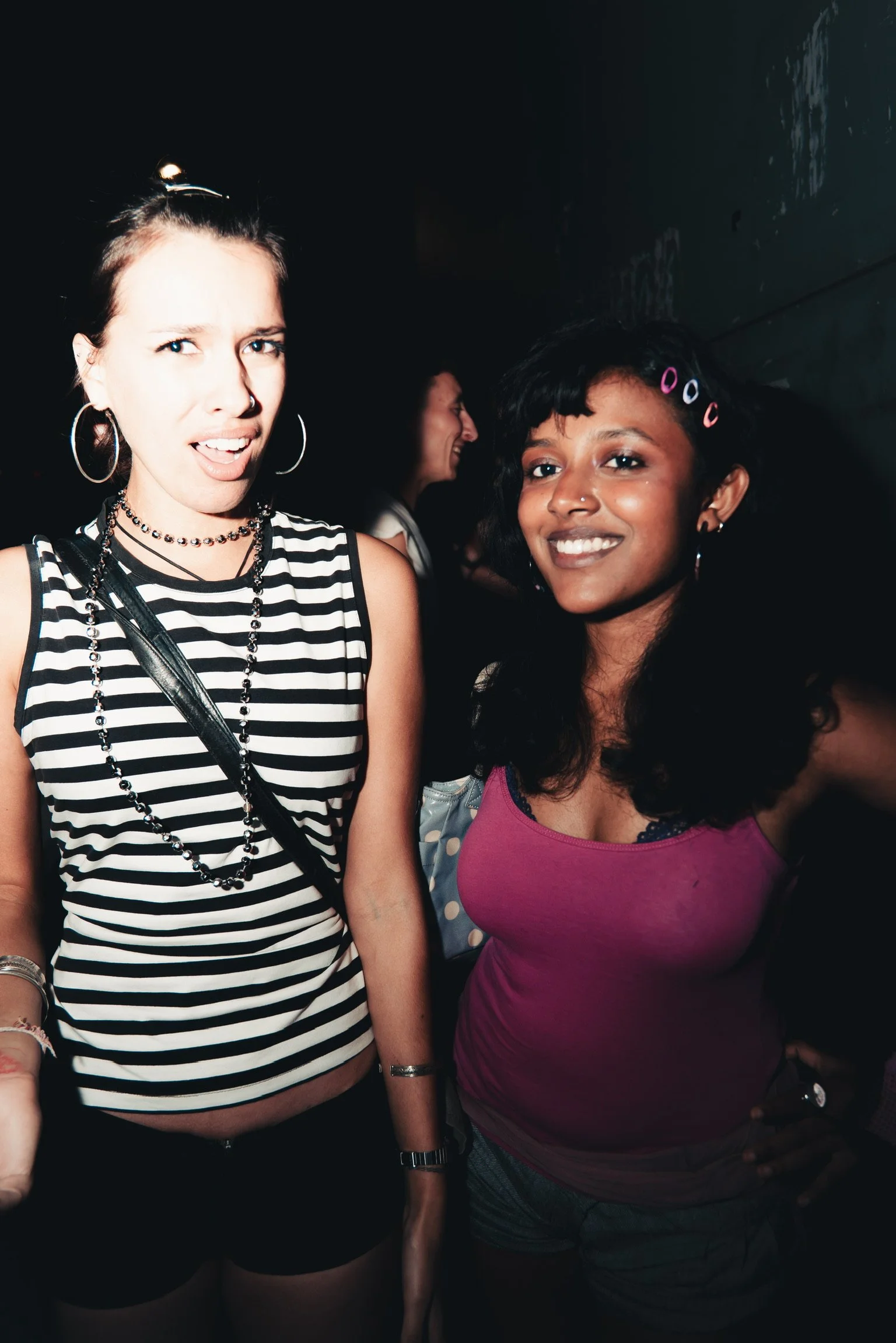Two women smiling at a social event, one wearing a black and white striped tank top and the other in a pink top with hair clips, with a dark background and a person in the background.