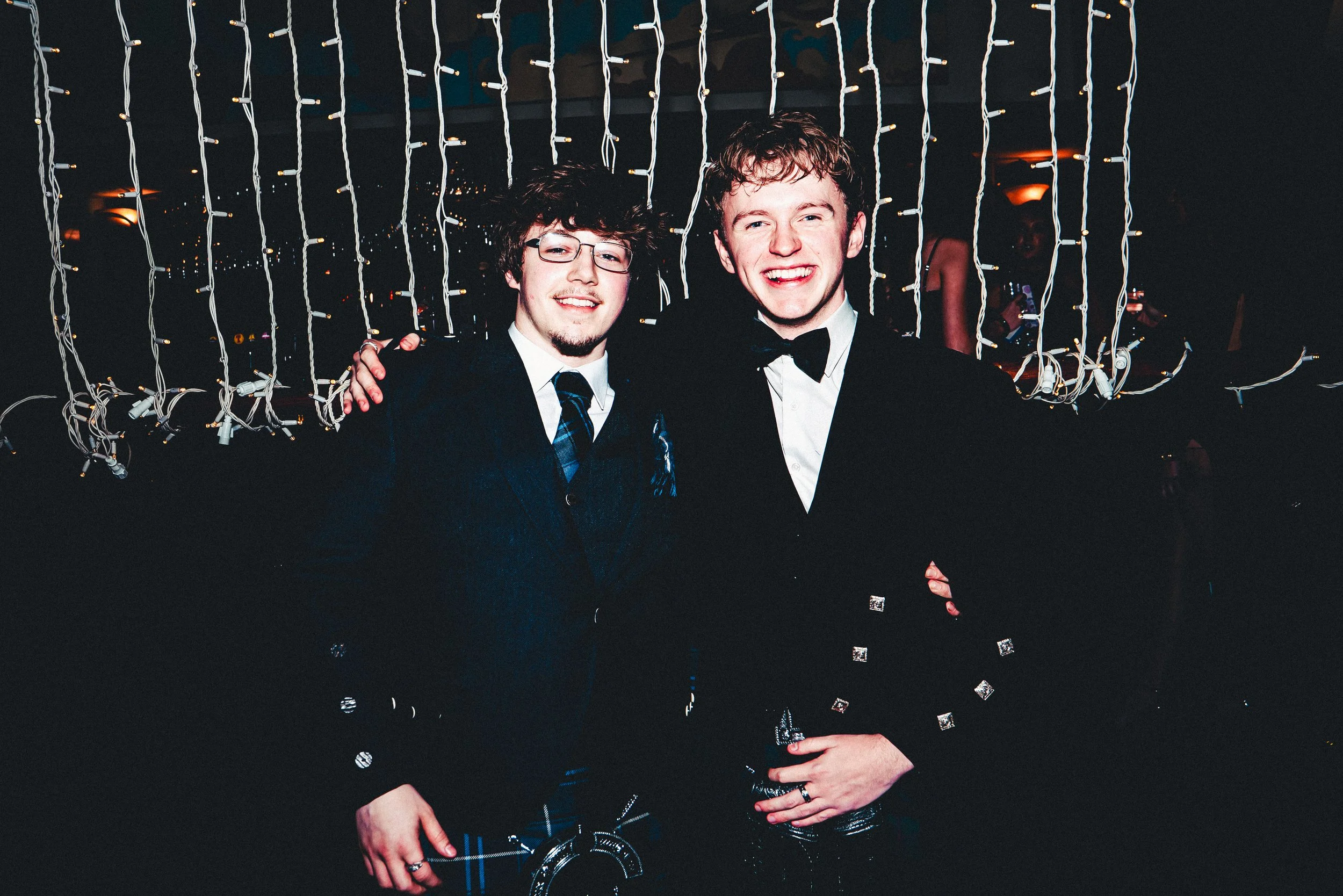Two young men dressed in tuxedos posing for a photo at a festive event, with holiday lights hanging behind them.