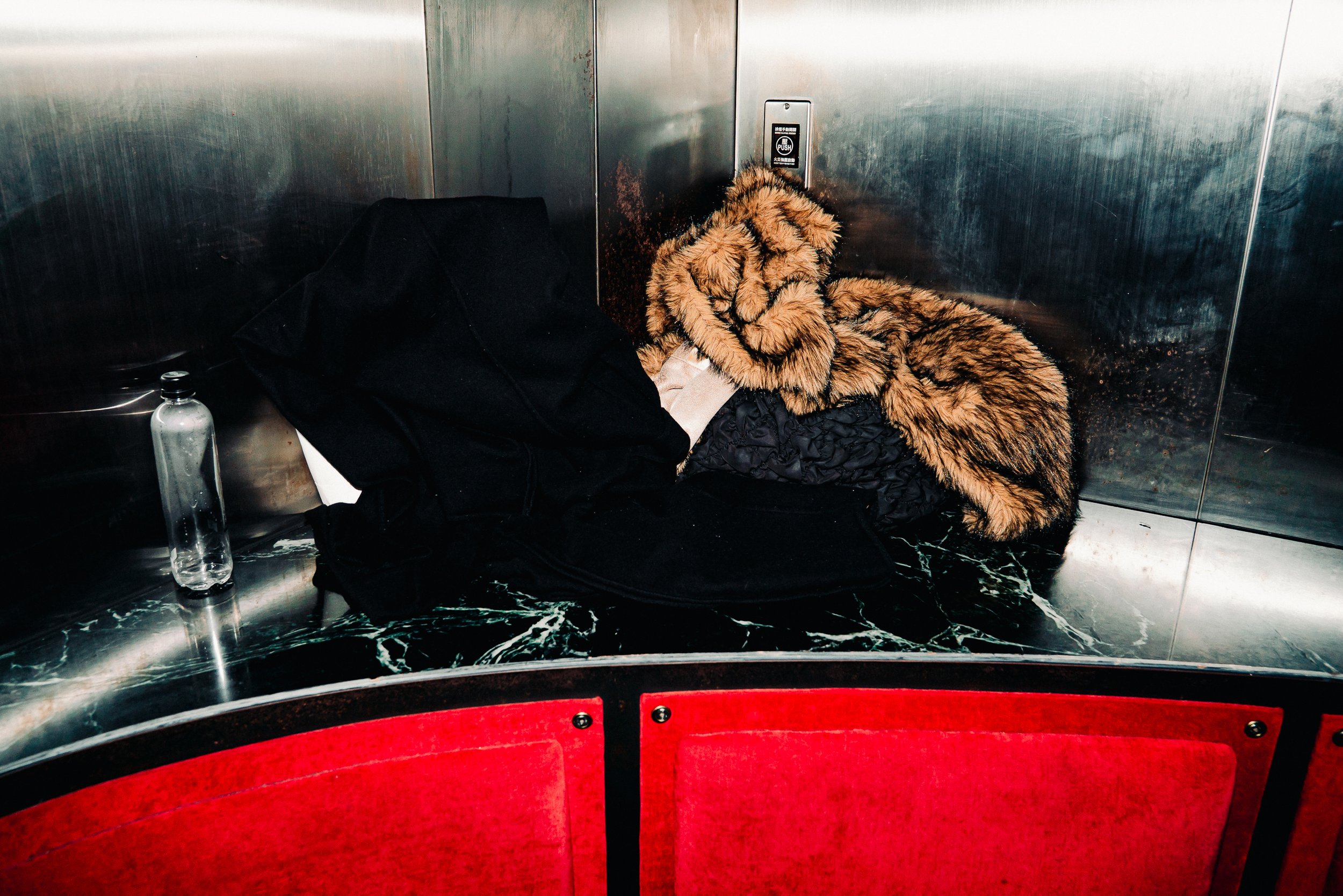 A person sleeping on the floor in an elevator, wearing a fur coat, surrounded by a black bag and a plastic water bottle, with red seats and metallic walls.