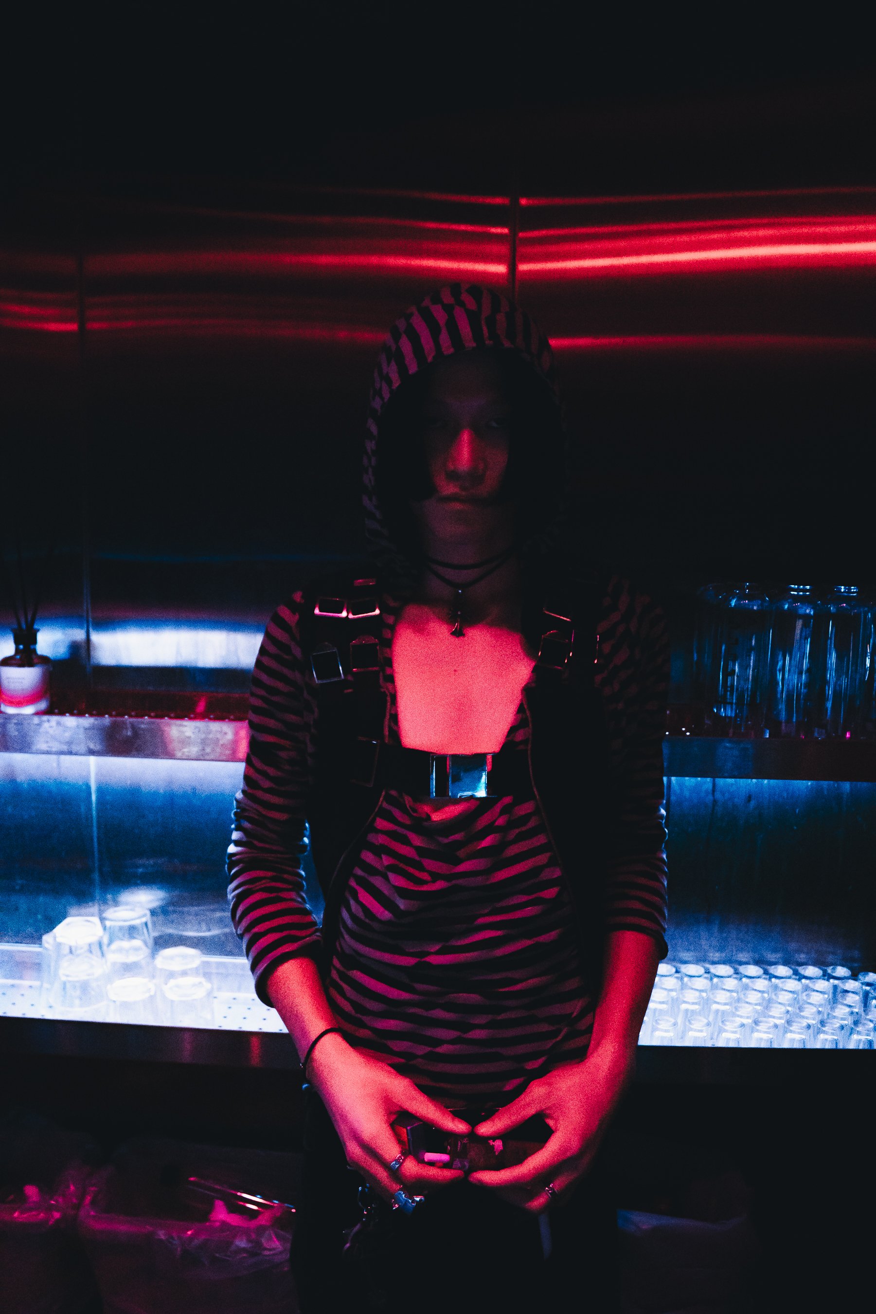 A person with dark hair wearing a striped hoodie with animal print, standing in a dimly lit bar or nightclub with red neon lights in the background. They are holding their phone with both hands.