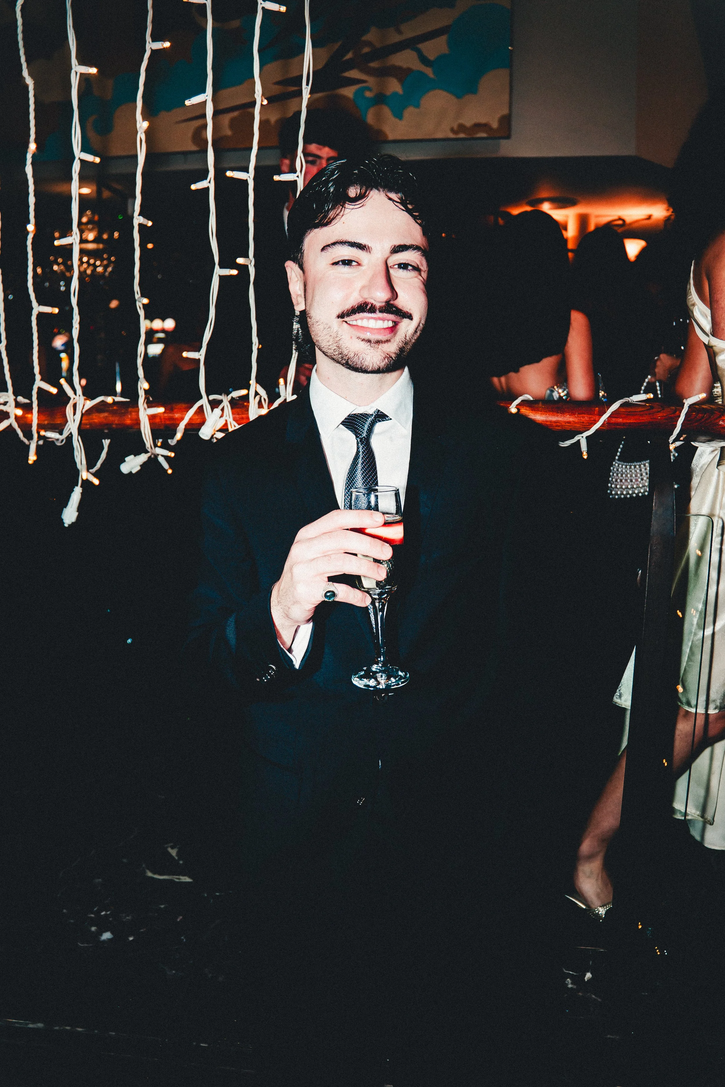 Young man in a tuxedo smiling and holding a drink at a party or nightclub. There are string lights and people in the background, with a woman wearing a shiny dress and heels.