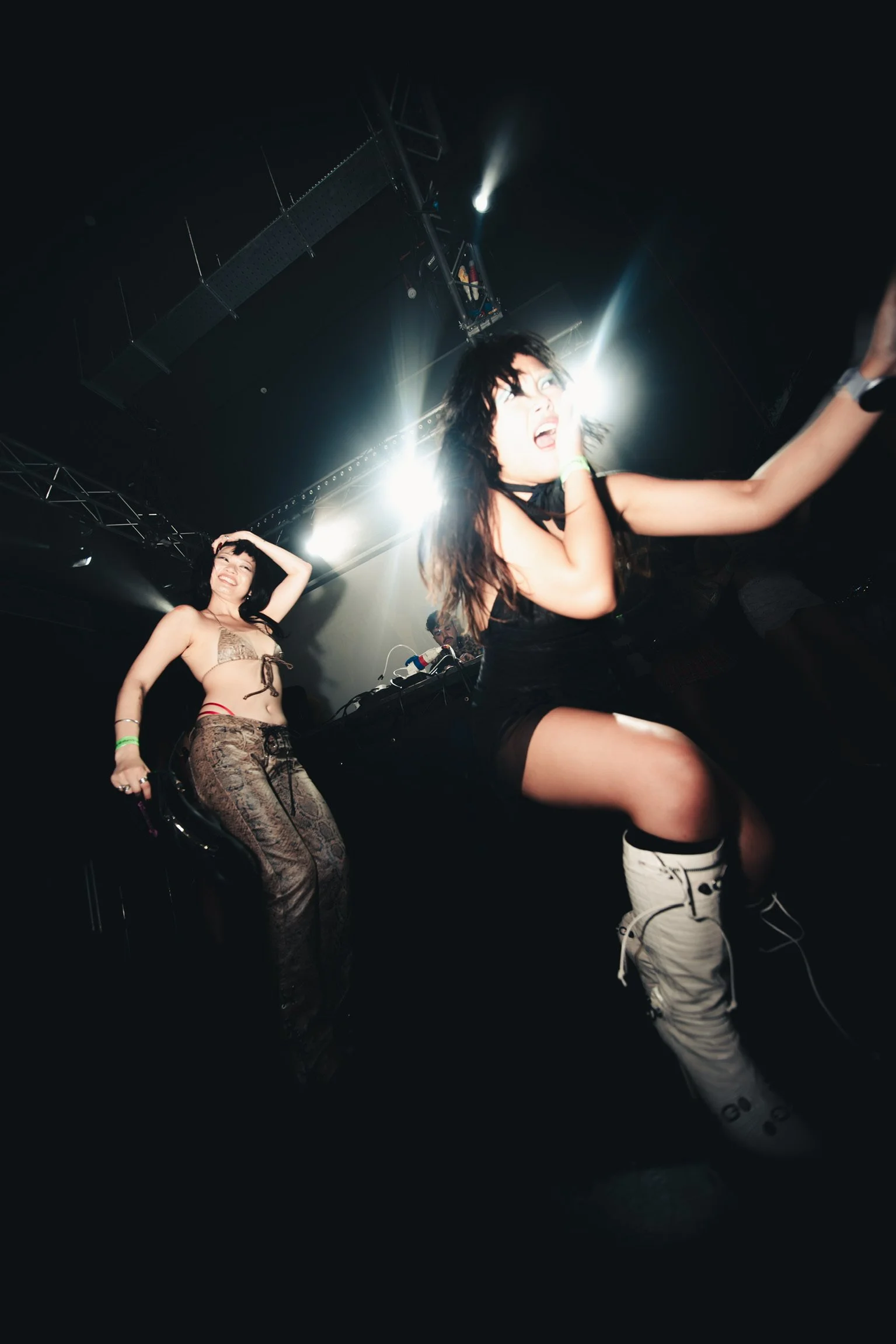Two women are at a nightclub or concert, one is wearing a black dress and white boots, and the other is wearing a beige patterned top and pants. They are dancing under bright stage lights with a DJ in the background.