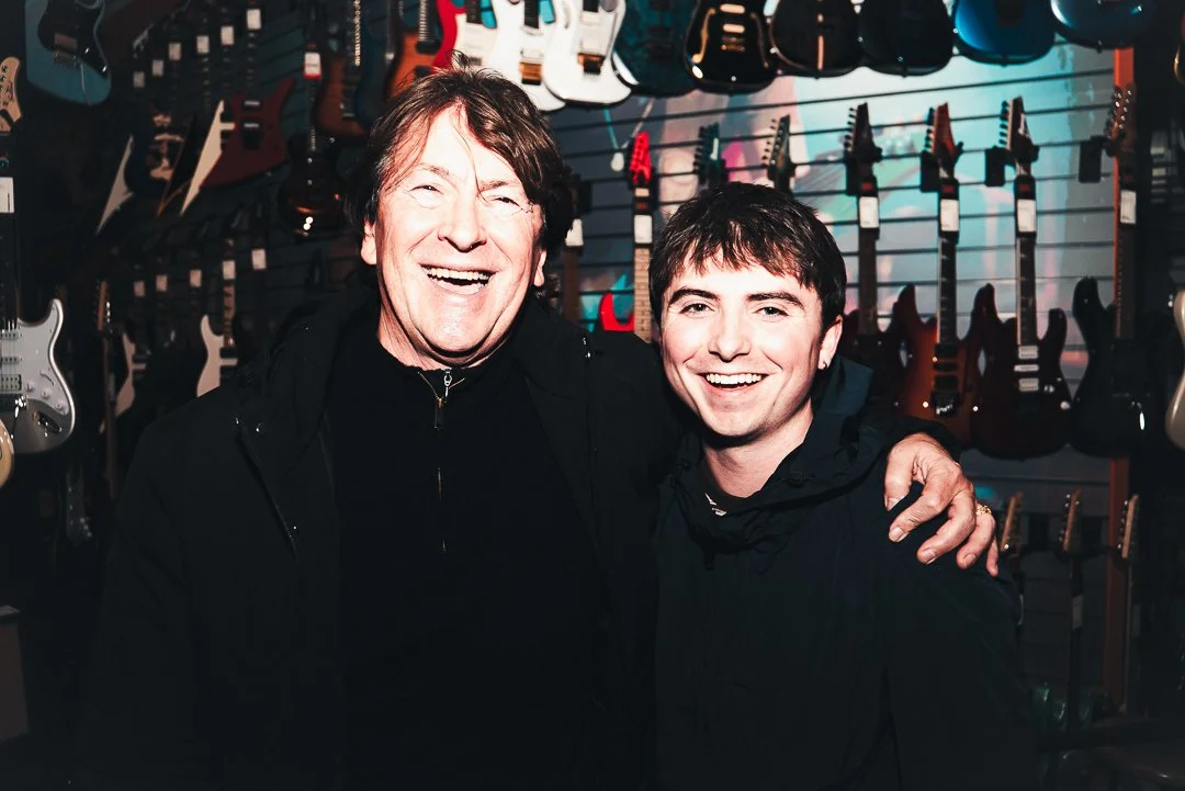 Two men smiling and posing in a guitar store with guitars hanging on the wall behind them.