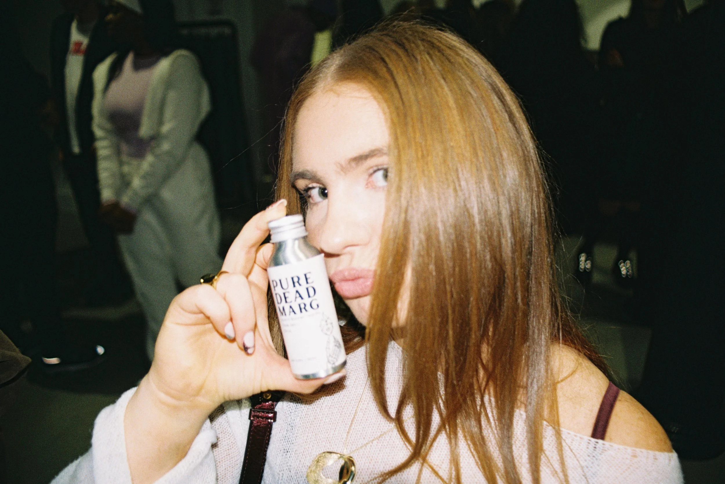 Young woman with red hair holding a small white bottle labeled 'Pure Dead Sea' near her eye, in a busy indoor setting.