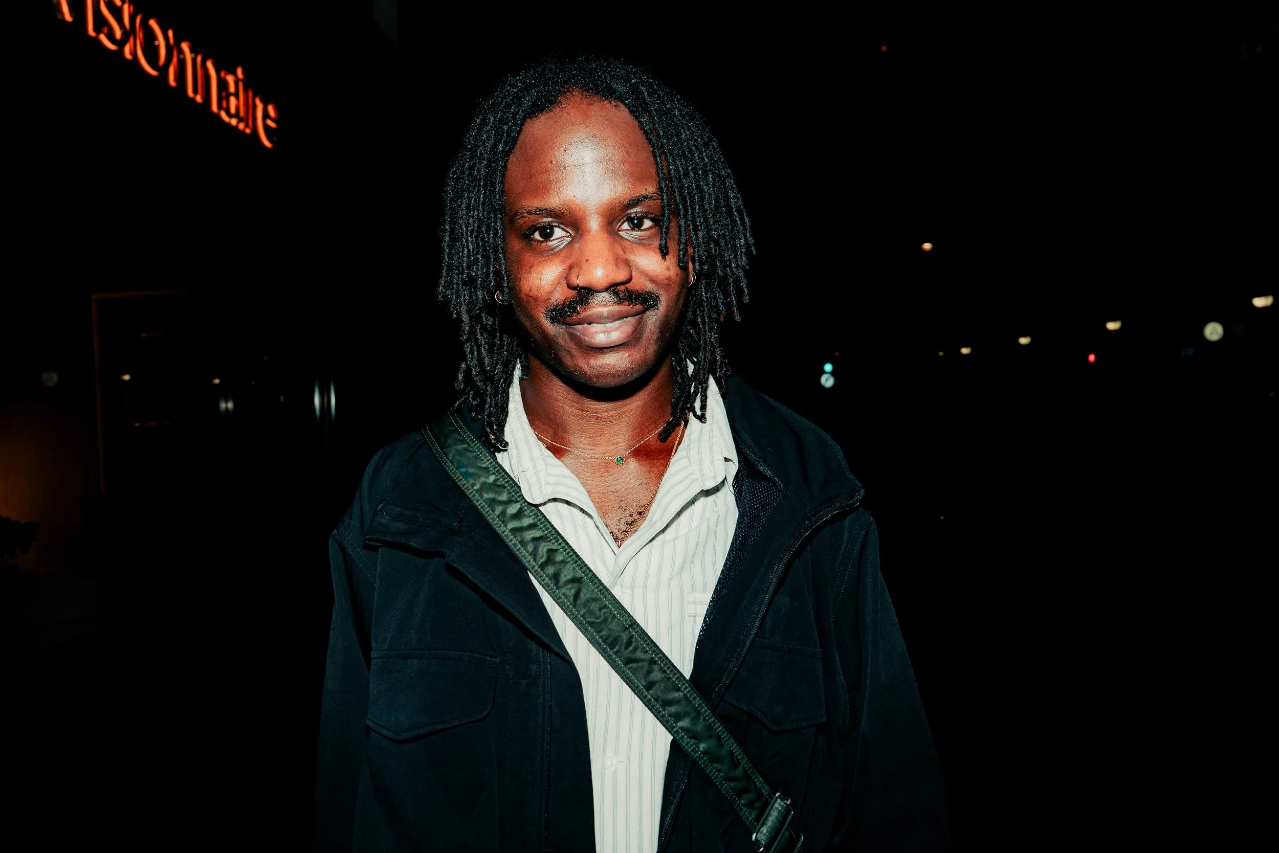 A person with dark skin and shoulder-length dreadlocks, wearing a nose ring, earrings, a striped shirt, and a dark jacket, standing outdoors at night with a neon sign in the background.