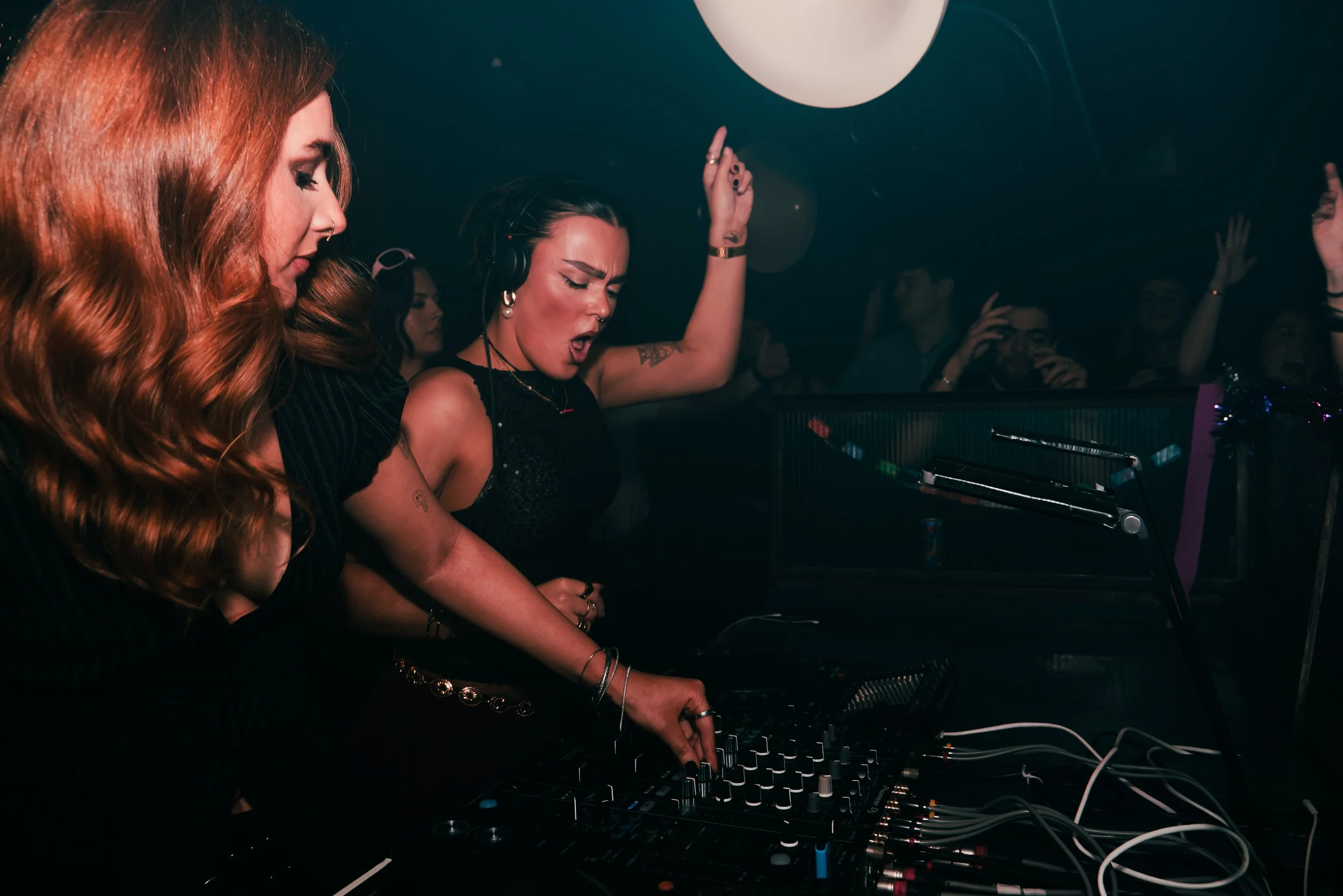 Two women DJing at a nightclub with a crowd enjoying the music in the background.