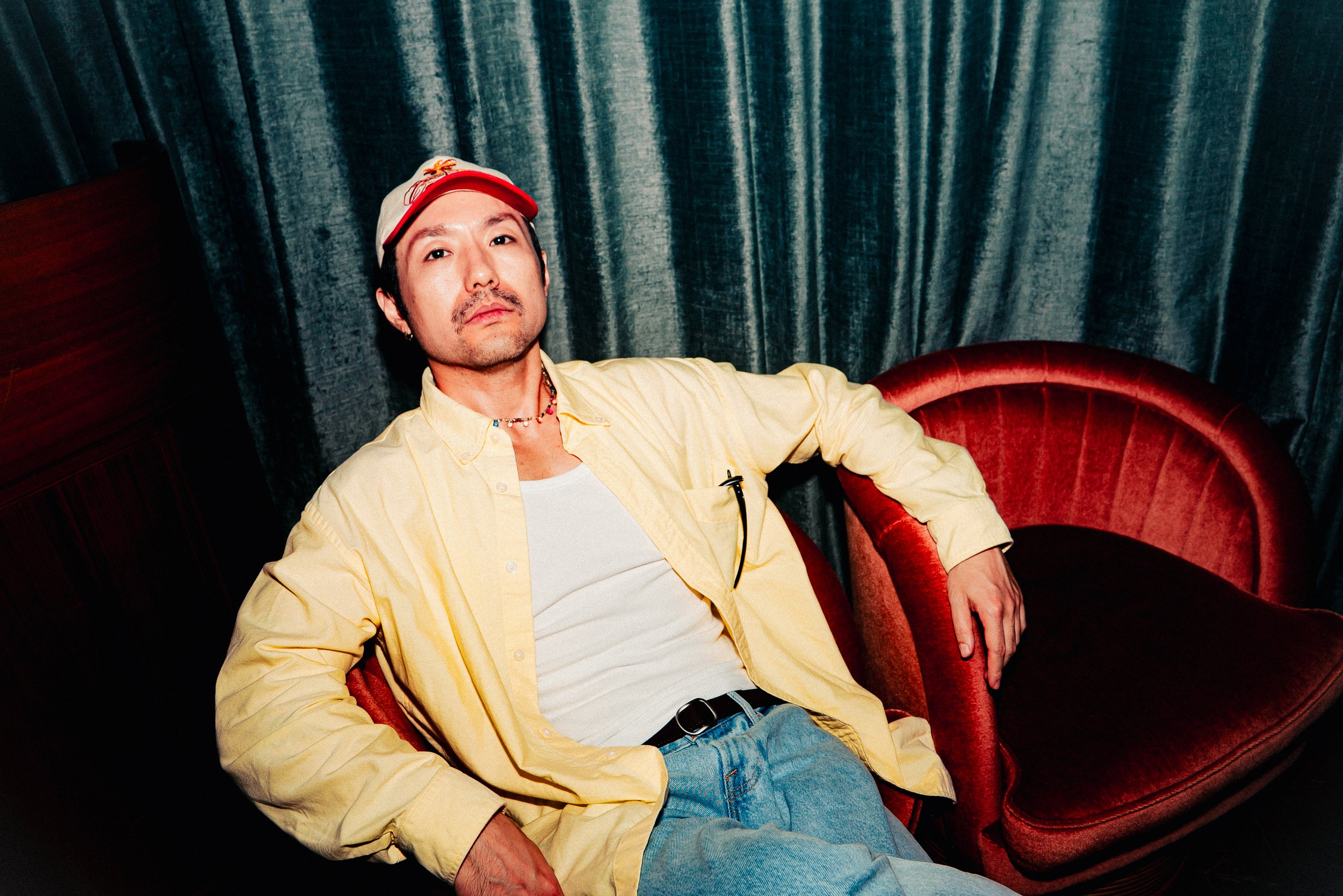A man with a mustache and beard, wearing a yellow shirt, white t-shirt, red cap, and jewelry, sitting on a red velvet armchair in front of a dark green velvet curtain.