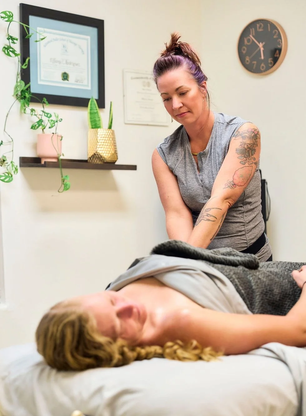 A  female massage therapist providing massage to a woman lying face down on a treatment table in an office setting with framed certificates, plants, and a clock on the wall.