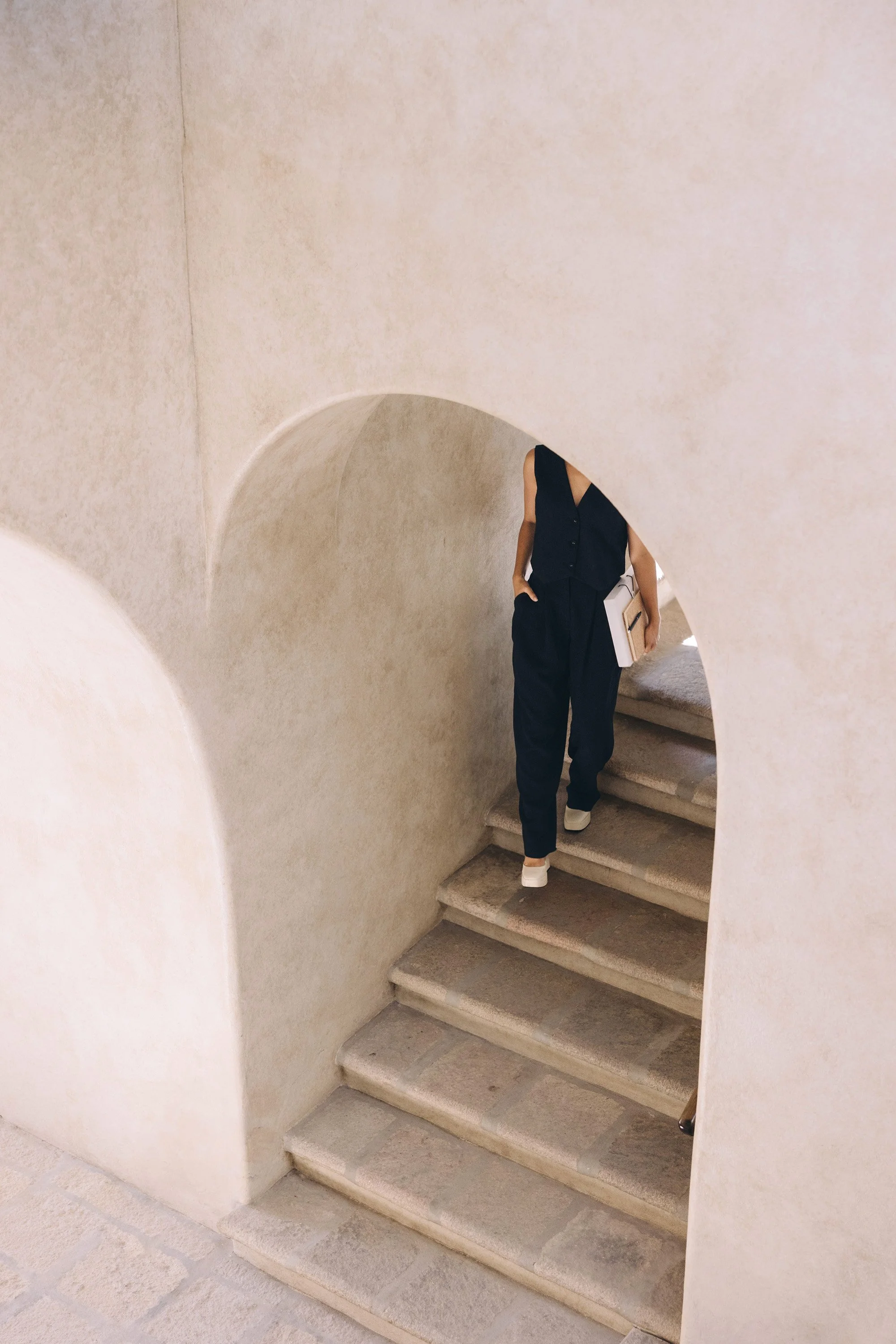 The Olive Branch VA. A person in dark clothing, carrying a folder or notebook, walking up a beige stone staircase through an arched passageway with beige textured walls.