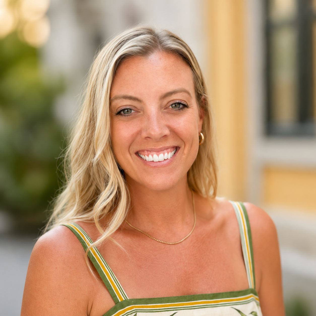 A smiling woman with blonde wavy hair wearing a sleeveless green, yellow, and white striped top standing outdoors with blurred background. The Olive Branch VA