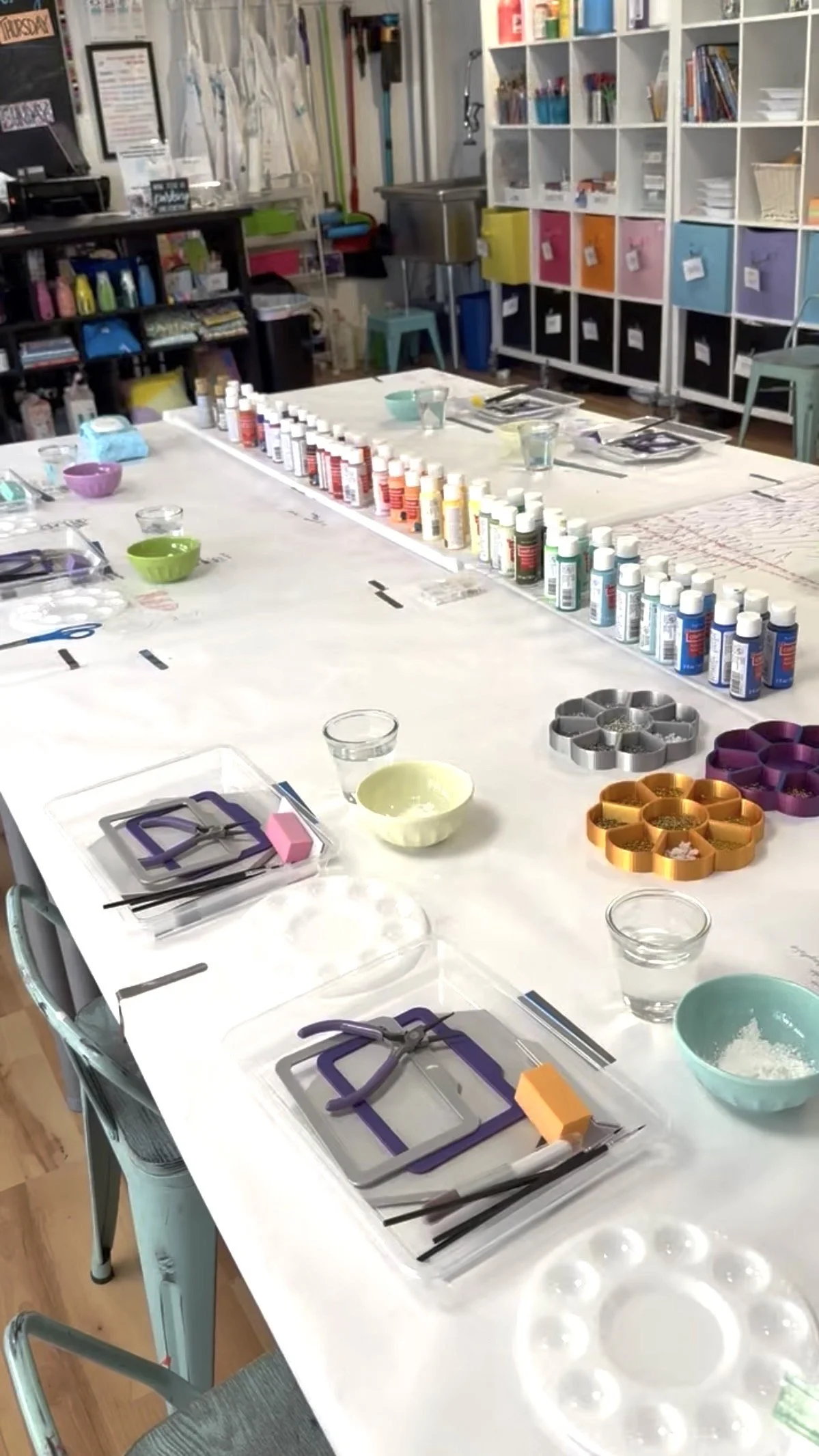 A craft workspace with various art supplies, including paint bottles, scissors, silicone molds, bowls, and tissue paper, set up on a white table in a classroom or studio.