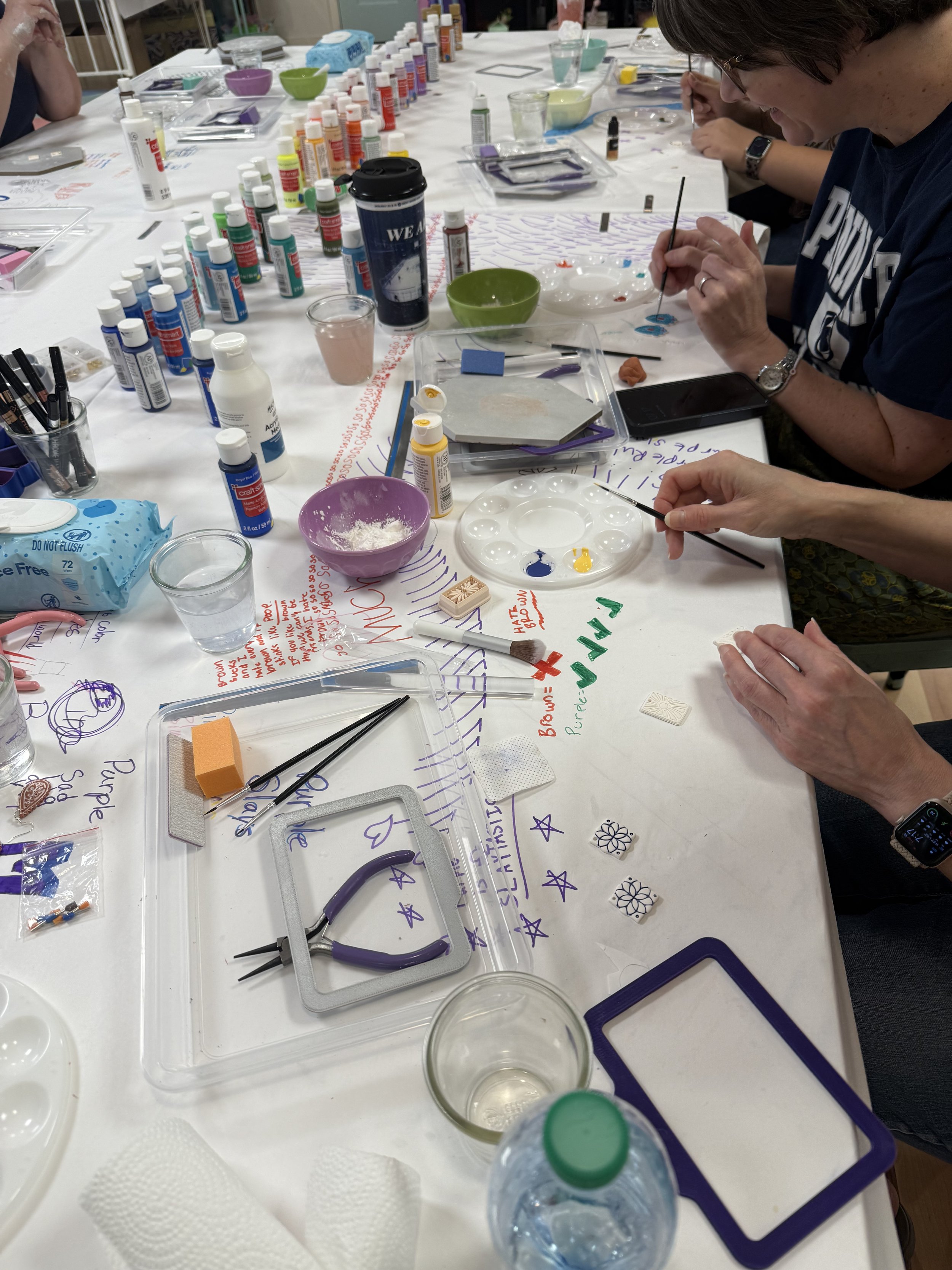 A table covered with various art supplies including paint bottles, paint palettes, brushes, paper towels, and glasses of water. People are painting with fine brushes, creating designs on paper.