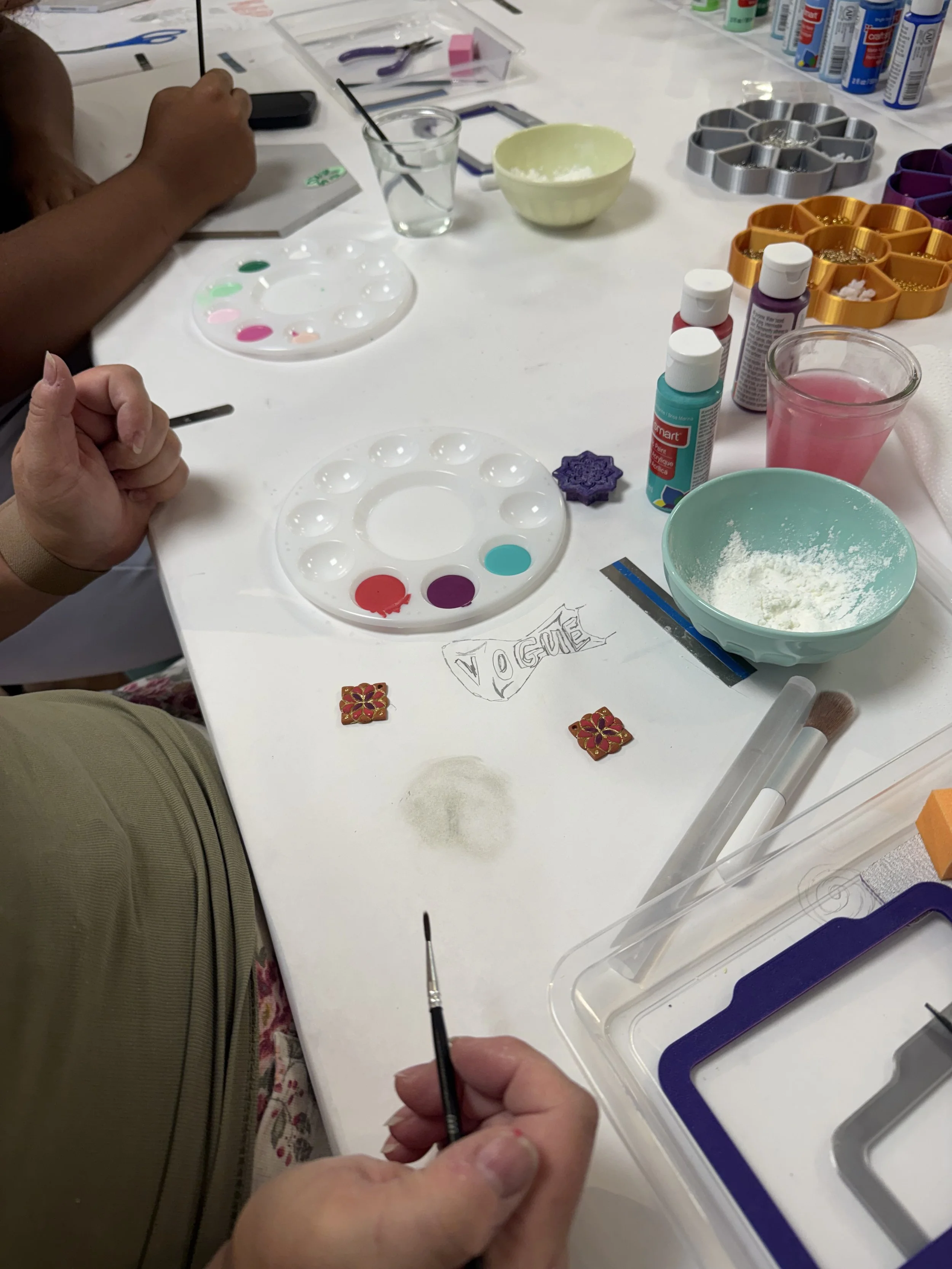 Person painting a decorative tile at a craft table, surrounded by painting supplies, tiles, and other craft materials.