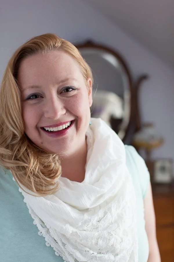 A smiling woman with blonde hair and light skin, wearing a white scarf and light blue top, standing in a room with a mirror and vintage furniture in the background.