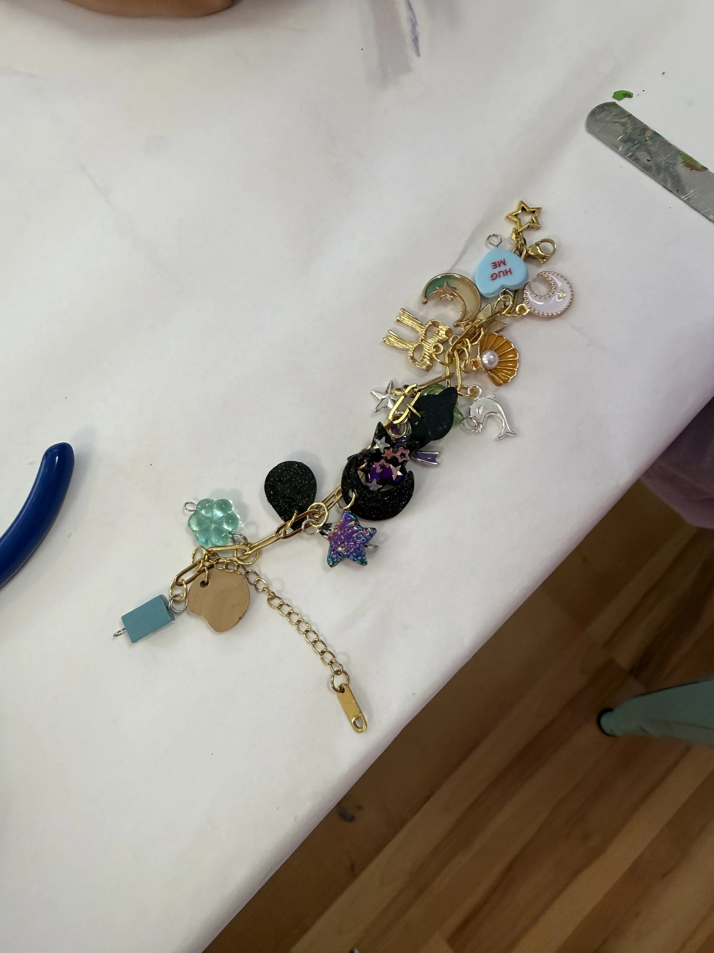 Assorted colorful charms and jewelry pieces scattered on a white surface, including stars, moon, and heart shapes, with a metal tool and a blue alligator clip nearby.