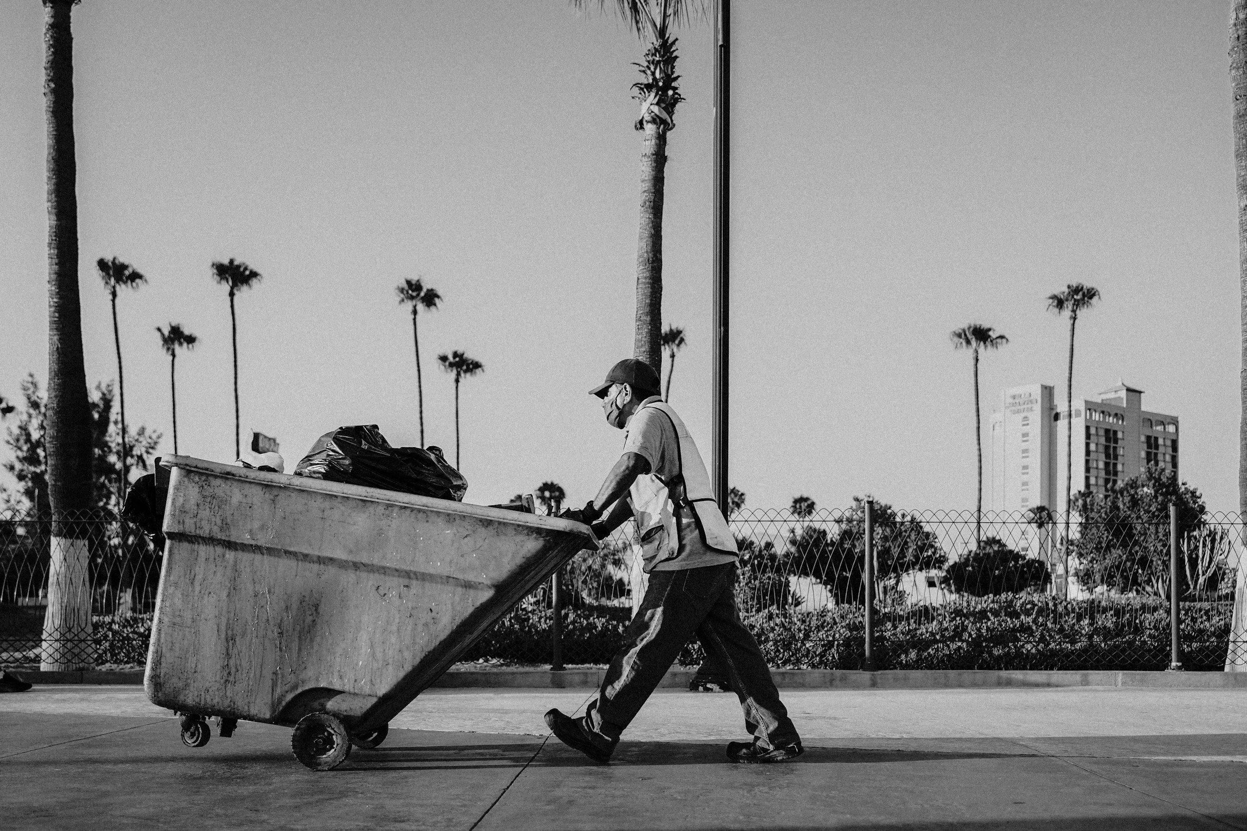 A person wearing a mask pushes a large garbage cart on a sidewalk. In the background, there are tall palm trees and buildings, indicating an urban area.