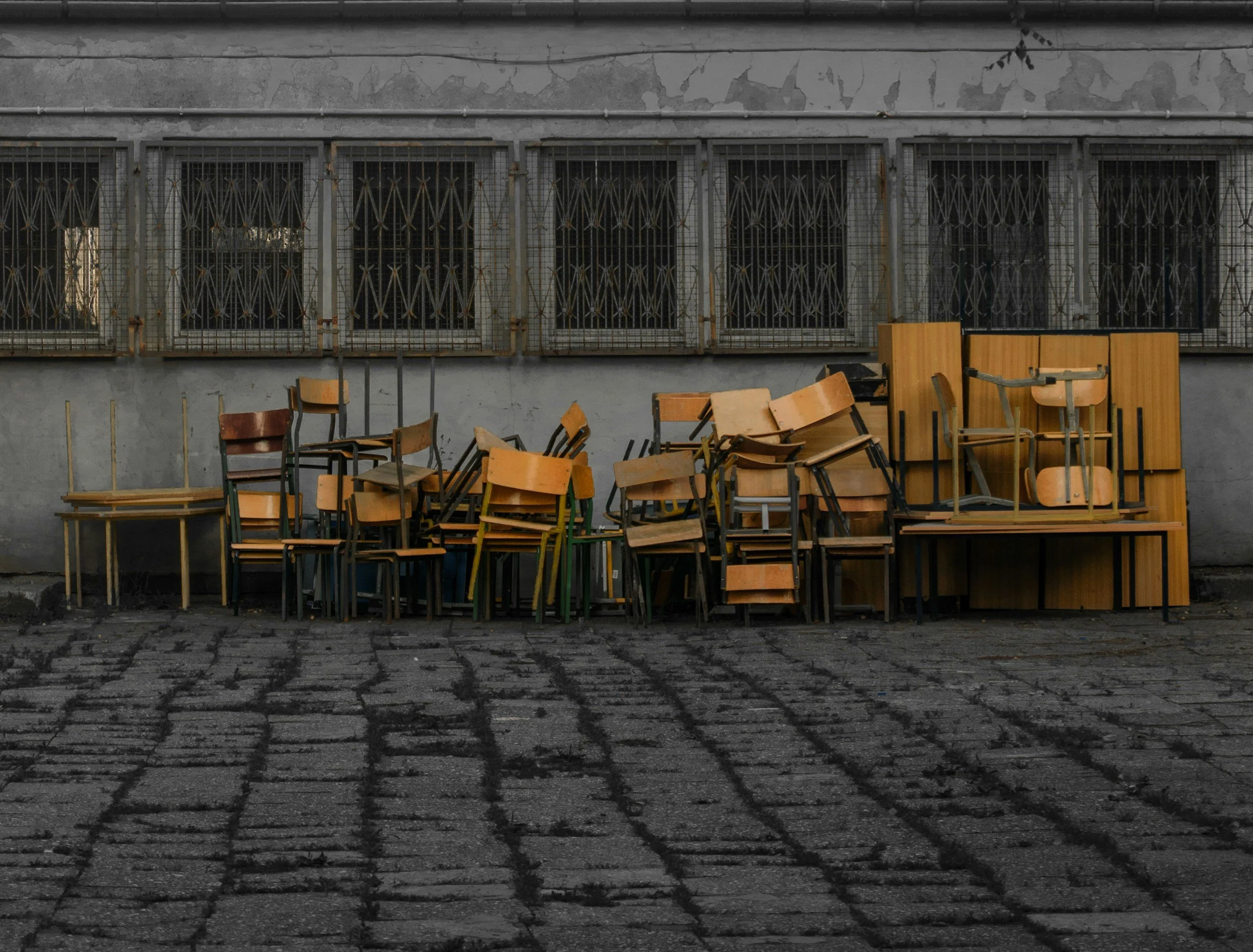 A row of old, rusted metal window grilles on a gray weathered wall, with scattered wooden chairs and tables in front, and a cobblestone street beneath.