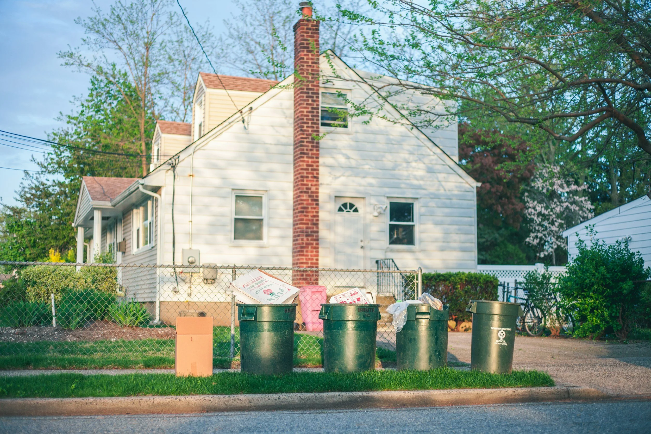 Trash cans in front of a chain-link fence with a house with a brick chimney and trees in the background.