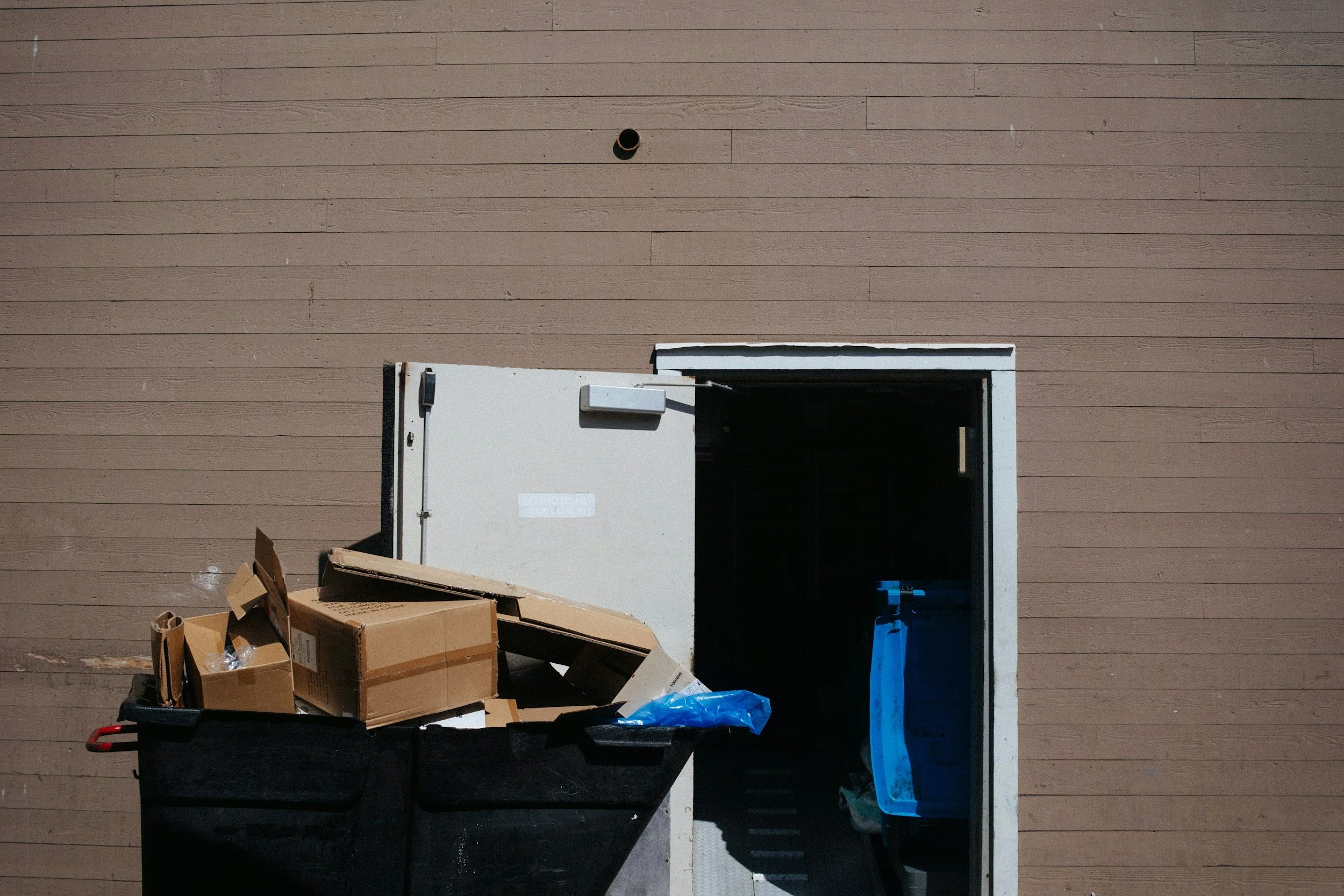A brown wooden wall with a small tunnel and an open door with a cardboard box outside. There are scattered cardboard boxes and trash in front of the wall, and blue plastic is visible.