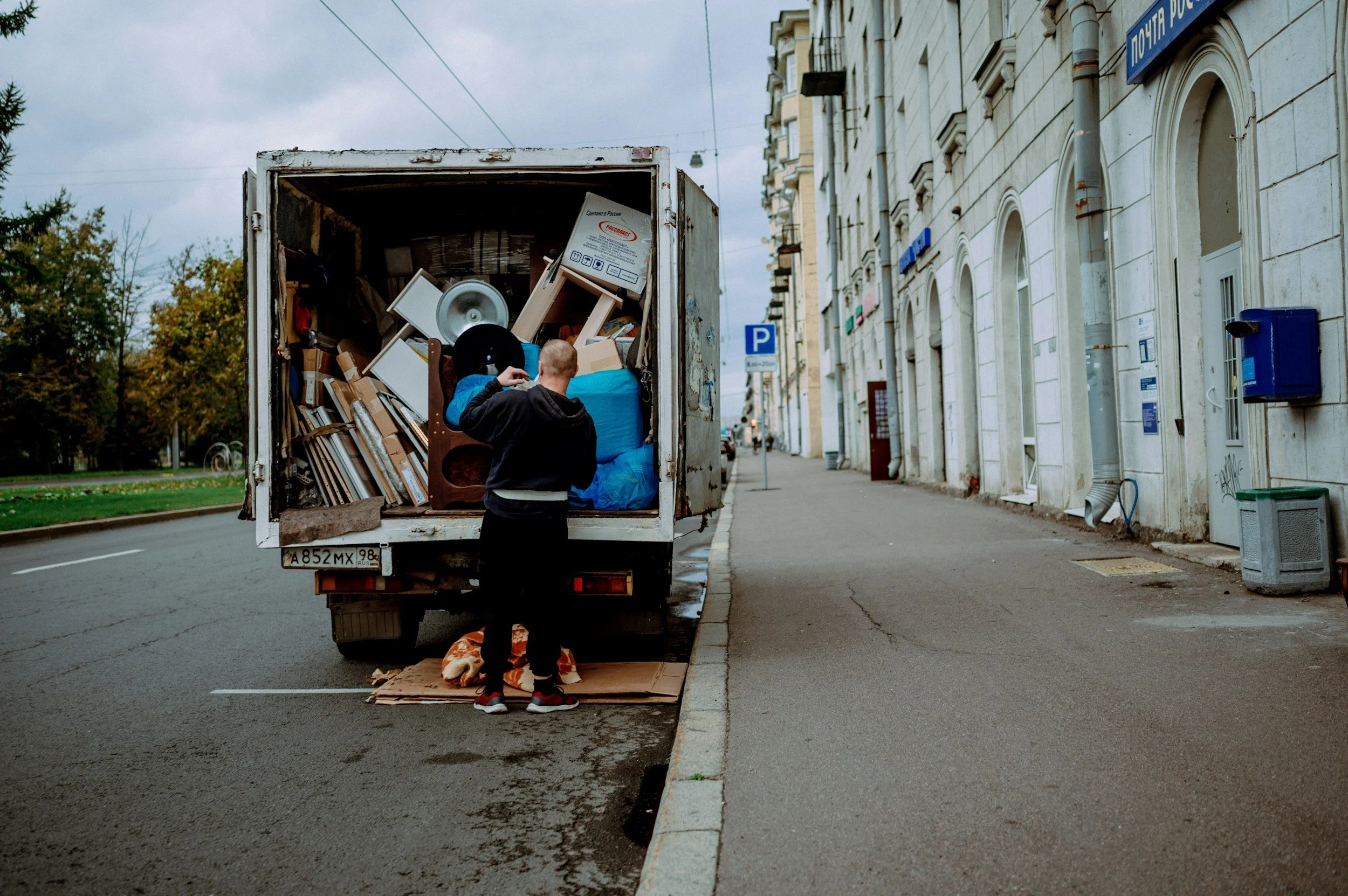 A person with a crew cut stands by a truck filled with furniture and boxes, with a bundle of cloth on the ground nearby. The scene takes place on a city street with buildings and trees in the background, overcast sky.