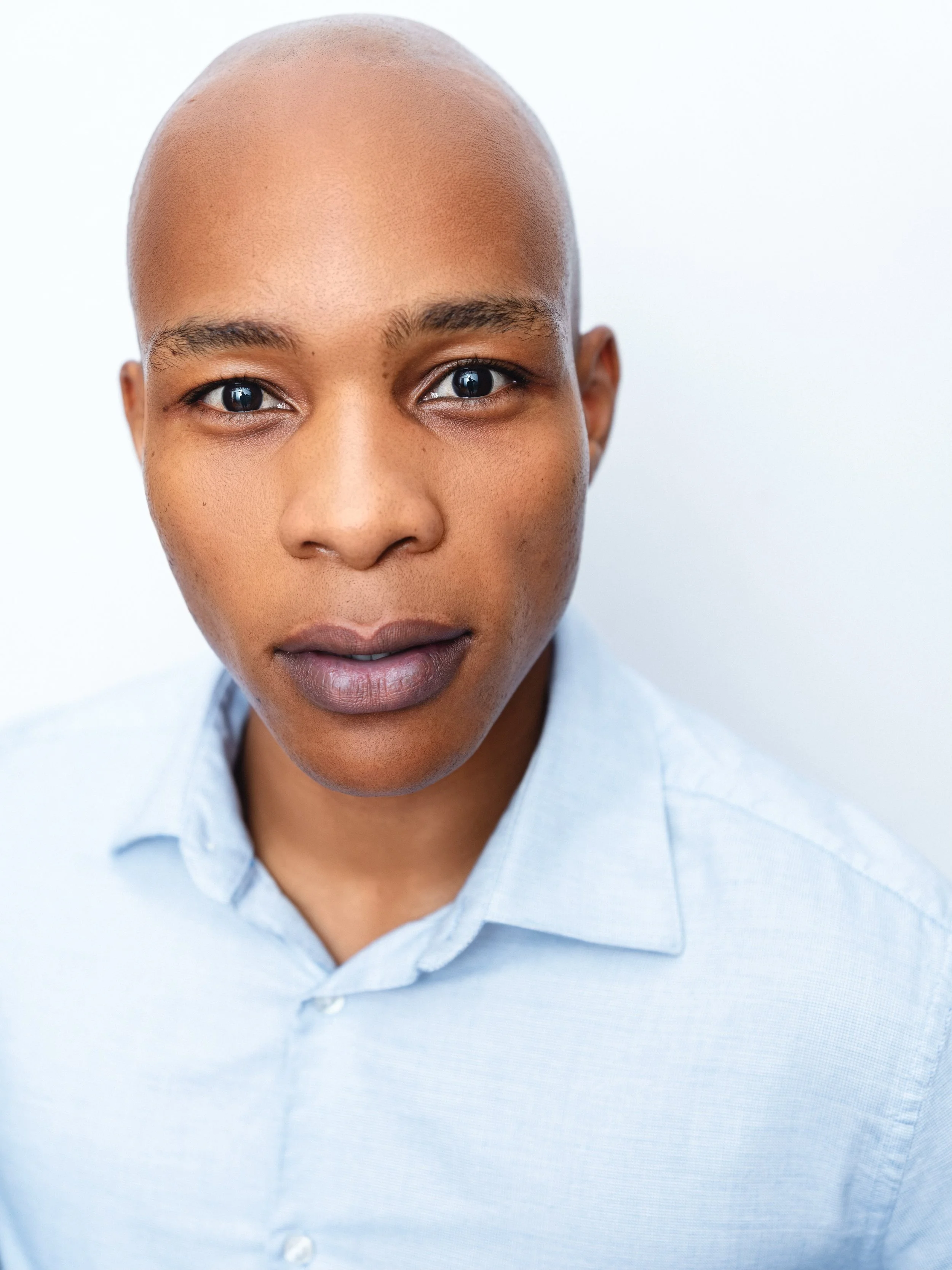 Close-up of a person's face showing a clean-shaven head and wearing a light blue collared shirt, against a plain light background.